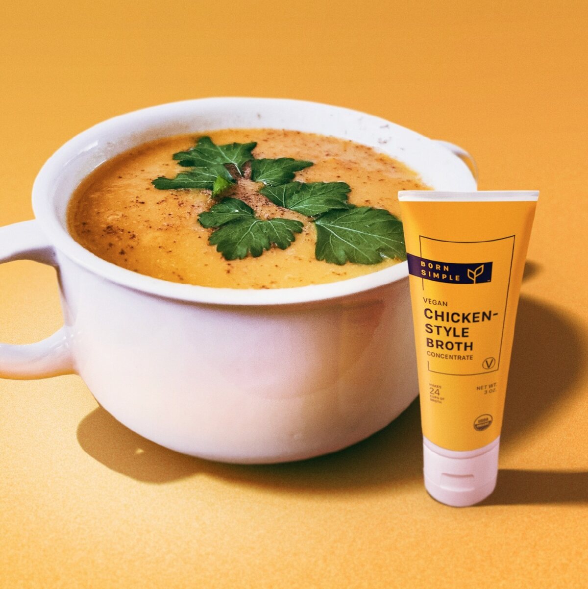 A white crockery bowl filled with autumn soup and next to an orange squeeze bottle of Born Simple vegan chicken-style broth against an orange background.