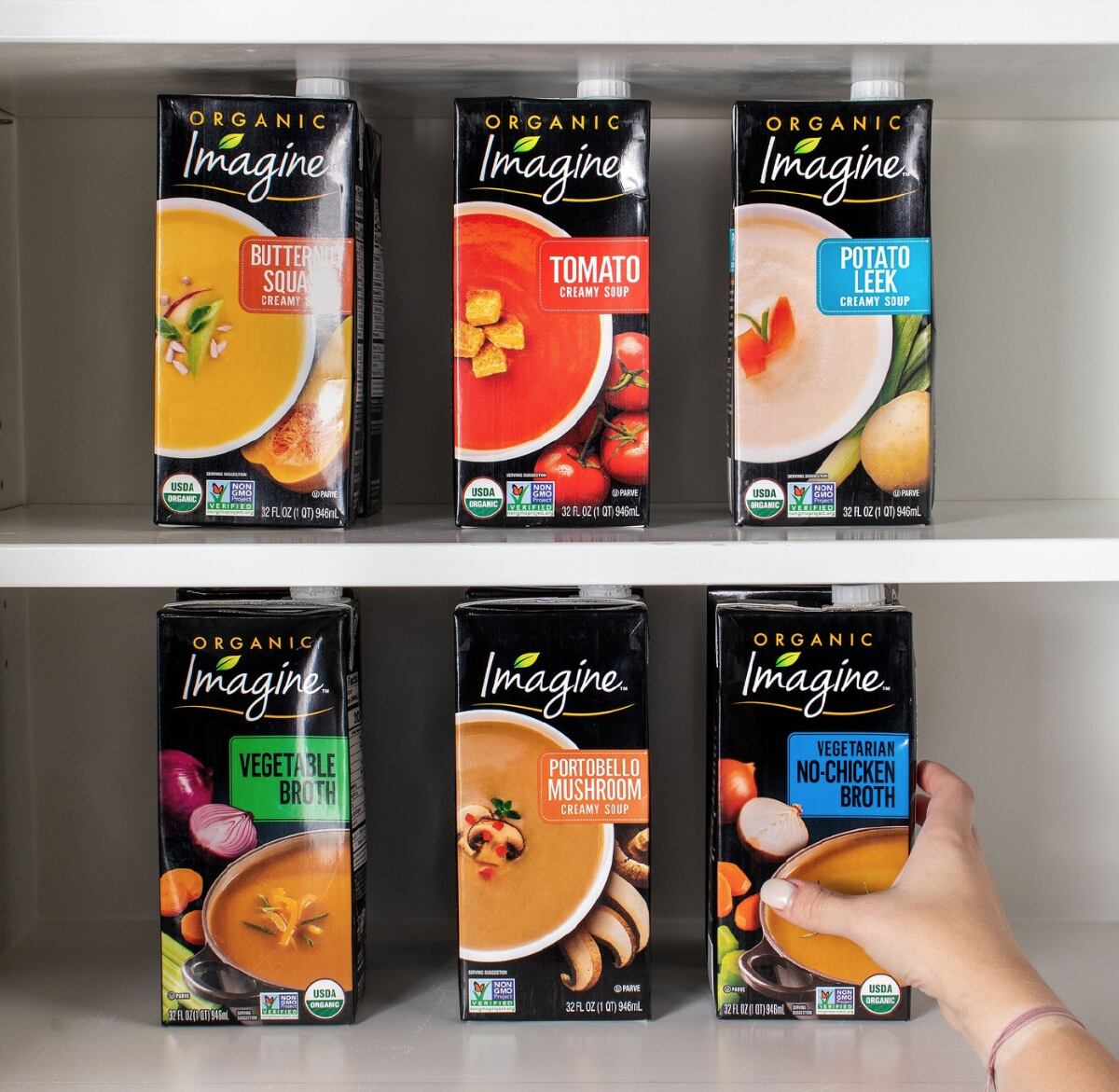 Six different flavors of Imagine soups and broths on two shelves of a kitchen cabinet.