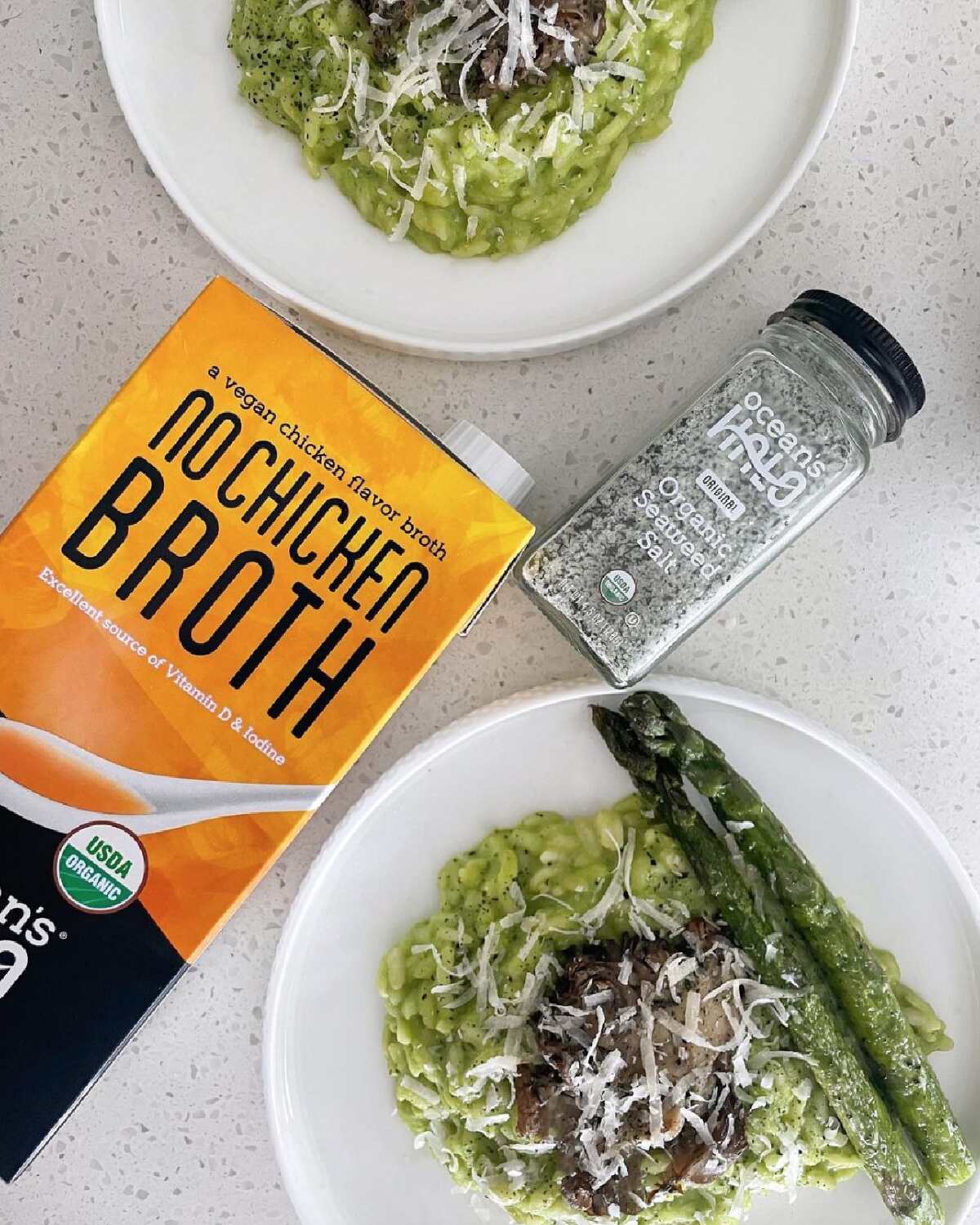 Two white plates full of veggie-filled food with asparagus next to an orange and black container of Ocean's Halo No Chicken Broth and a glass jar of seaweed salt. 