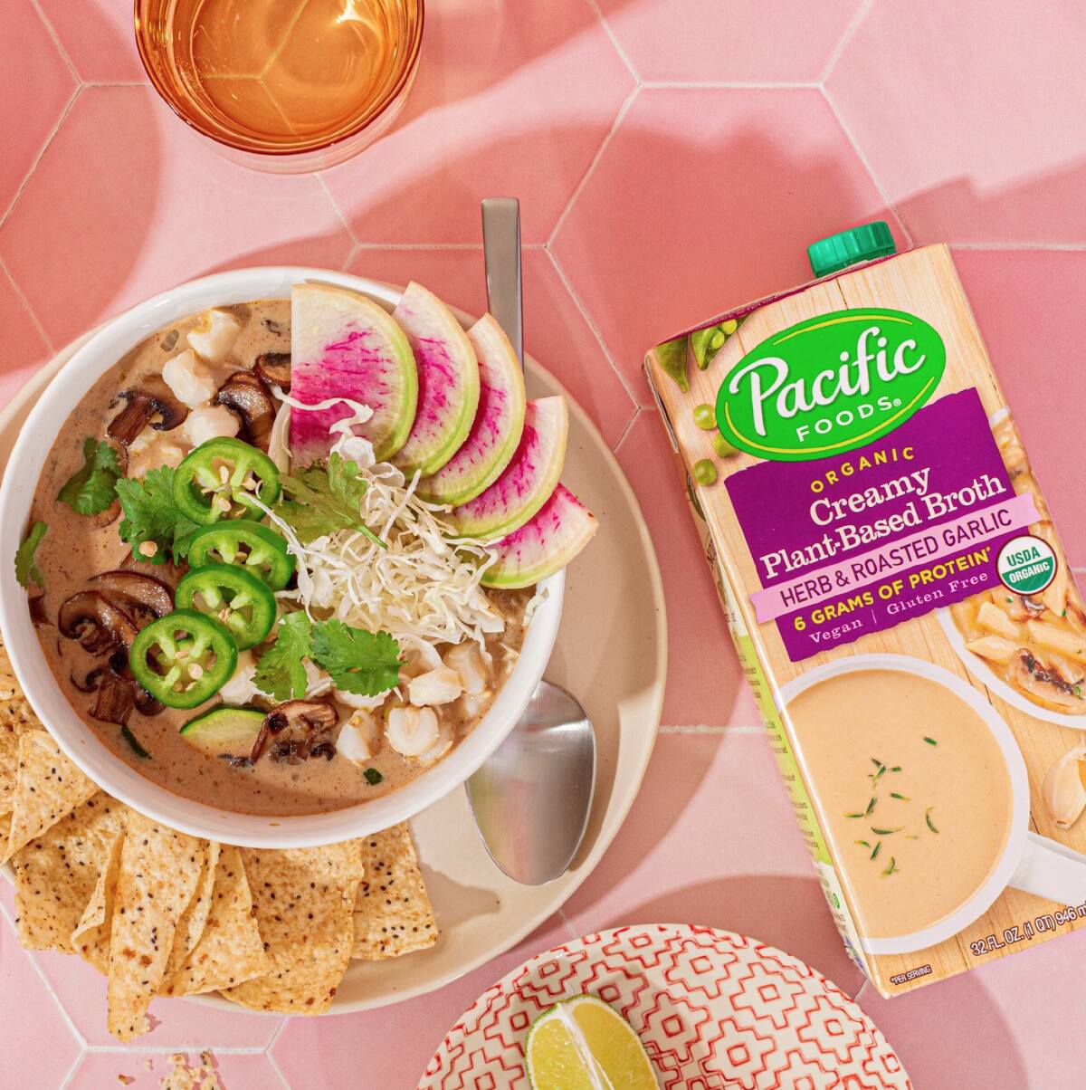 A bowl of warm soup and tortilla chips next to a container of Pacific Foods Plant-Based Herb & Roasted Garlic Broth against a pink background.