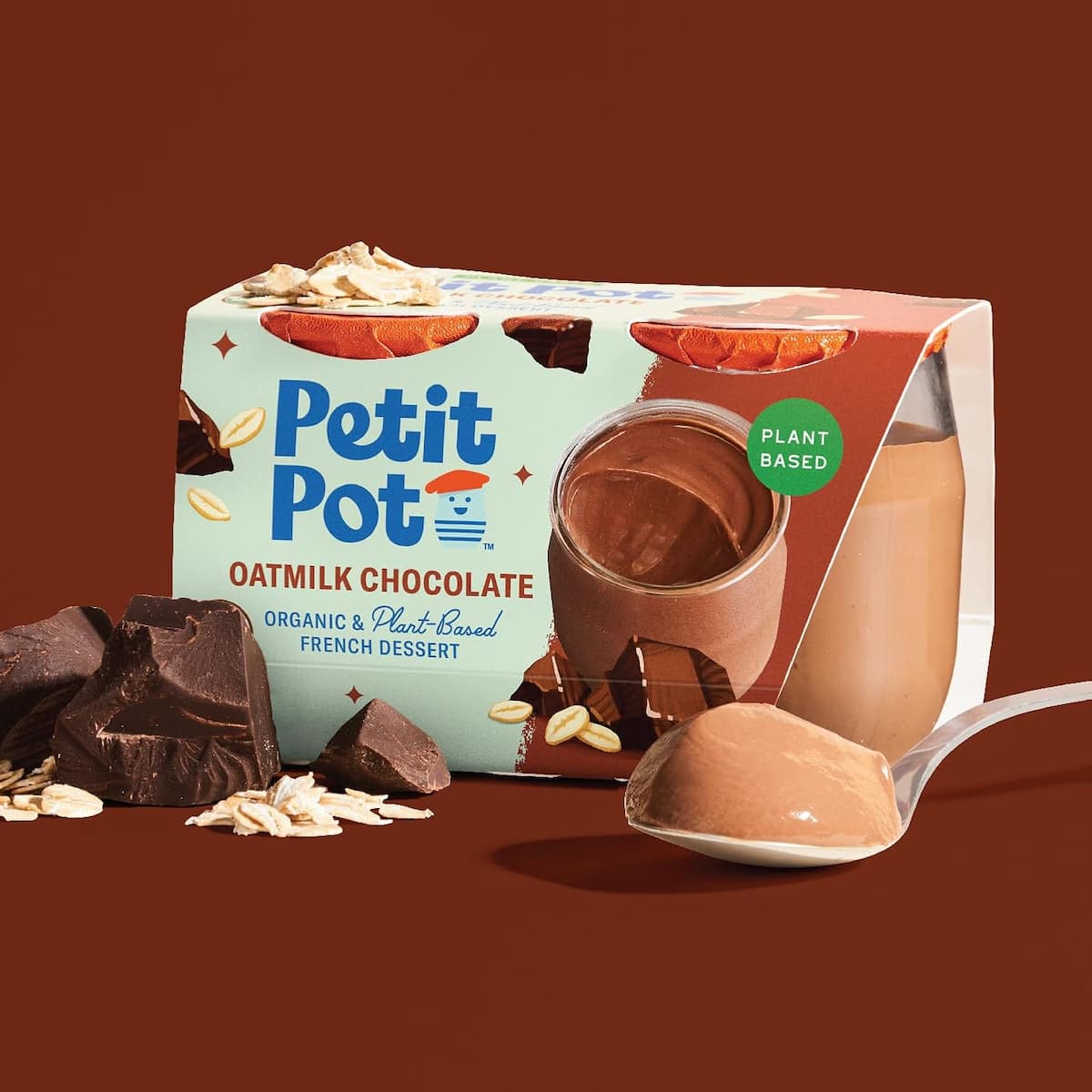 A two-pack of organic oatmilk chocolate pudding by Petit Pot next to a spoonful of pudding, oats and chocolate on a brown background.