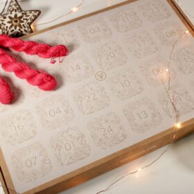 A craft box decorated and cut with advent day boxes accessorized with red skeins of yarn and fairy lights.