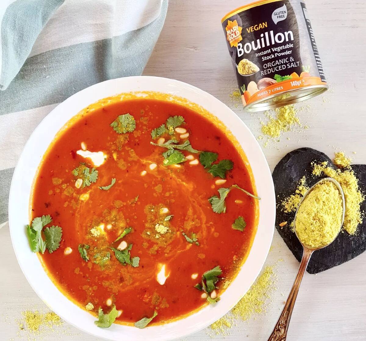 A white bowl filled with tomato soup and herbs next to a can of Marigold vegan vegetable bouillon and a spoon with the power still in it.