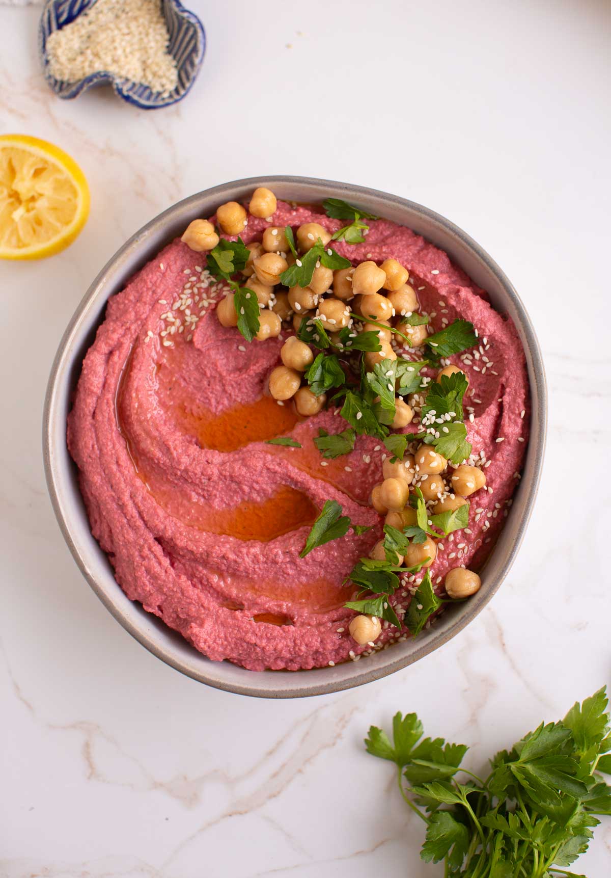 A bowl of pink beet hummus with decorative toppings served surrounded by ingredient flaylay. 