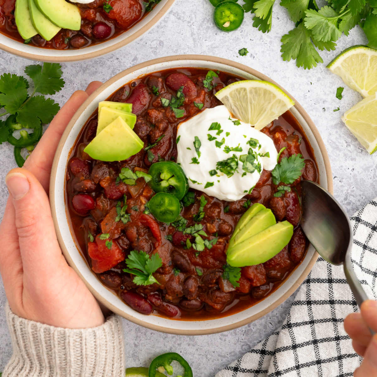 A bowl of vegan chili topped with fresh herbs, vegan sour cream, avocado slices, and a hand with a spoon about to scoop some up.