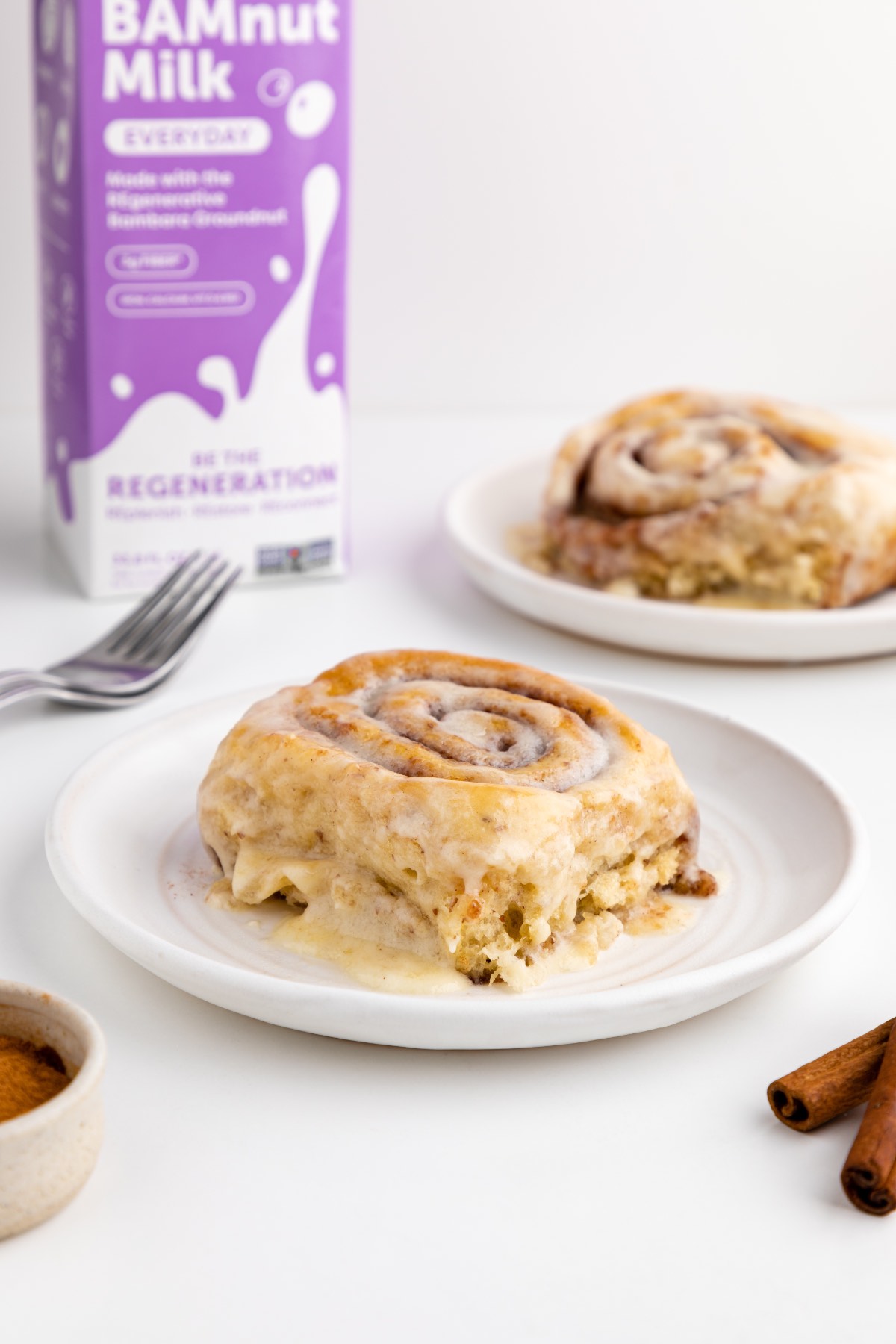 A ooey-gooey vegan cinnamon roll served on a white plate with cinnamon sticks next to it. 