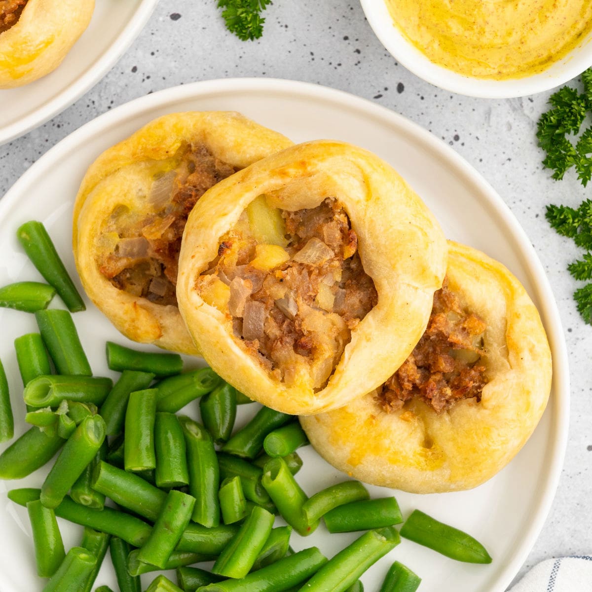 Three knishes on a plate served with green beans and mustard.