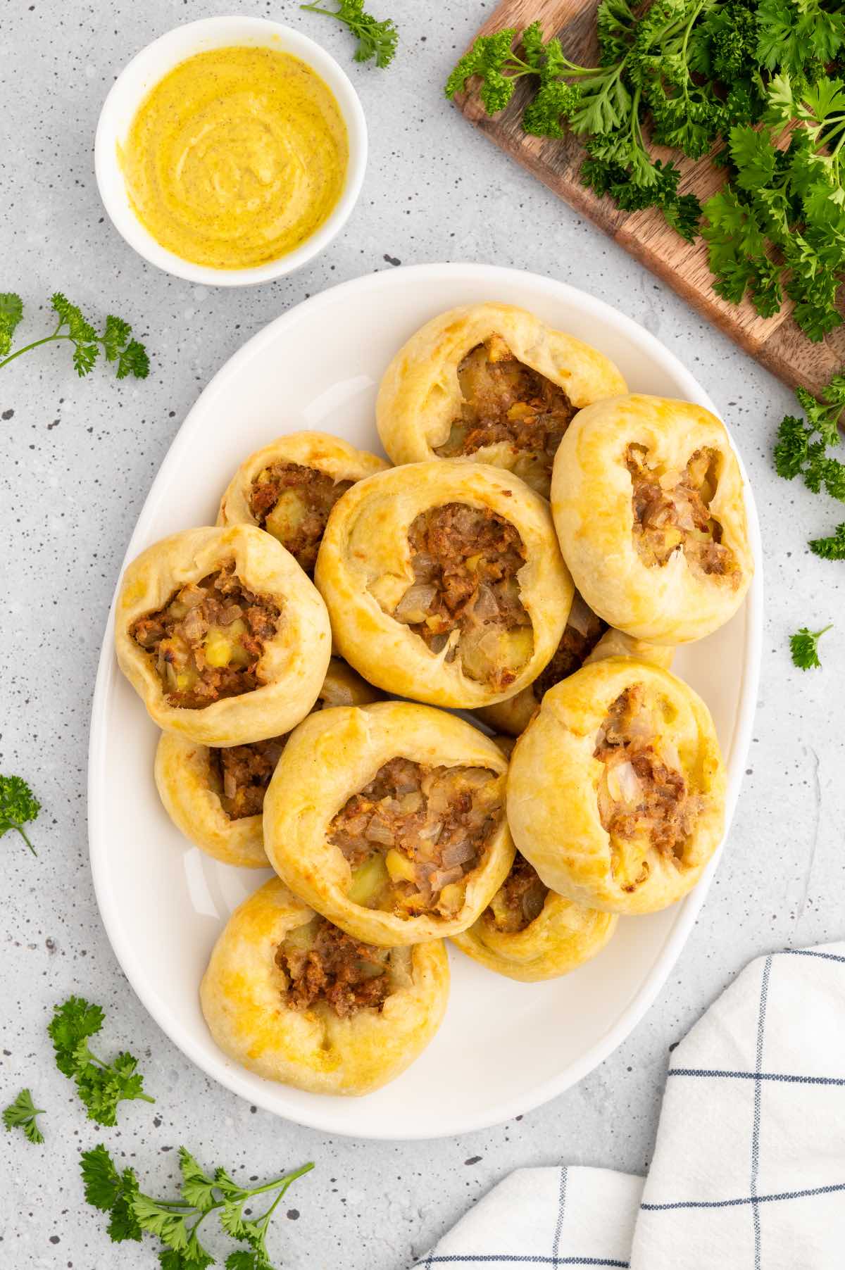 A white serving platter piled with vegan potato knish pastries garnished with parsley and served with a side of mustard. 