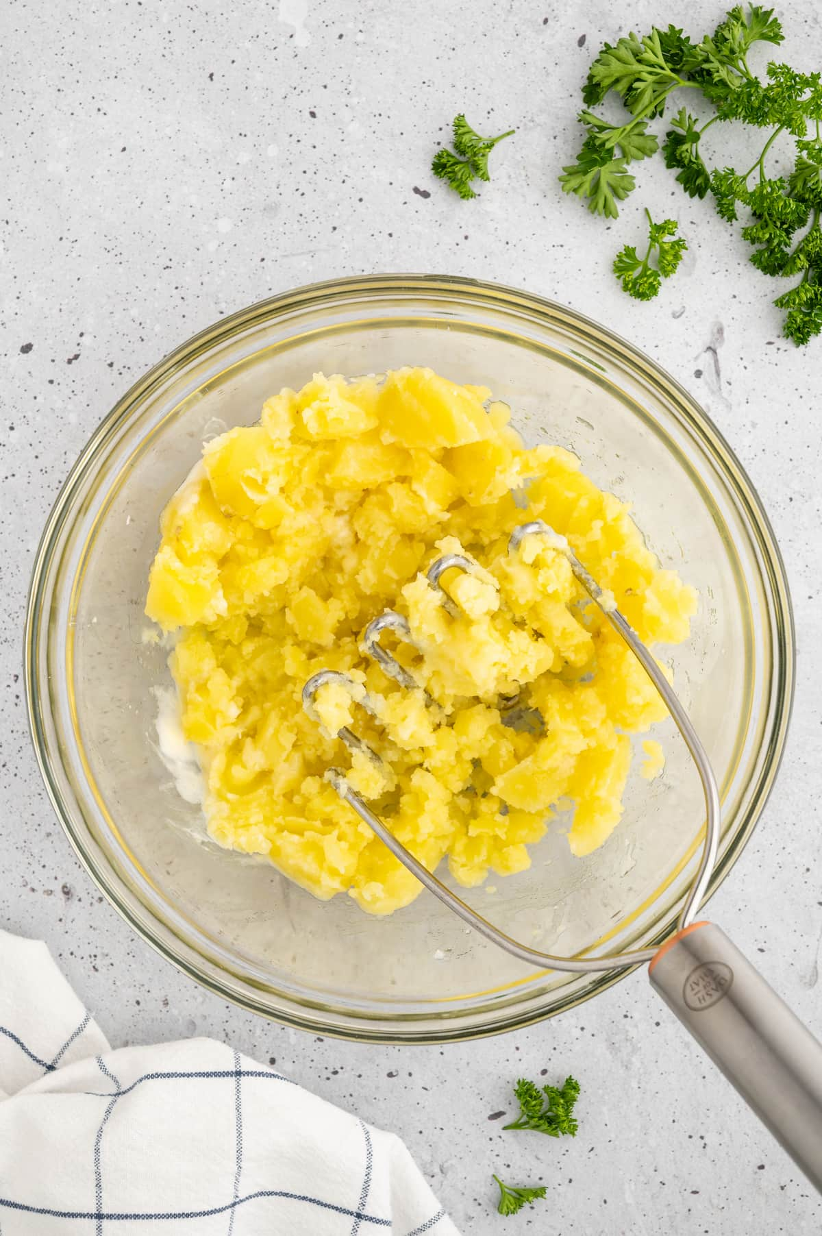 A glass bowl with yukon gold potatoes being mashed into mashed potatoes using a potato masher.