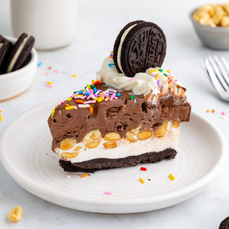 A slice of vegan ice cream cake on a plate, topped with vegan sprinkles, buttercream, and a vegan sandwich cookie.