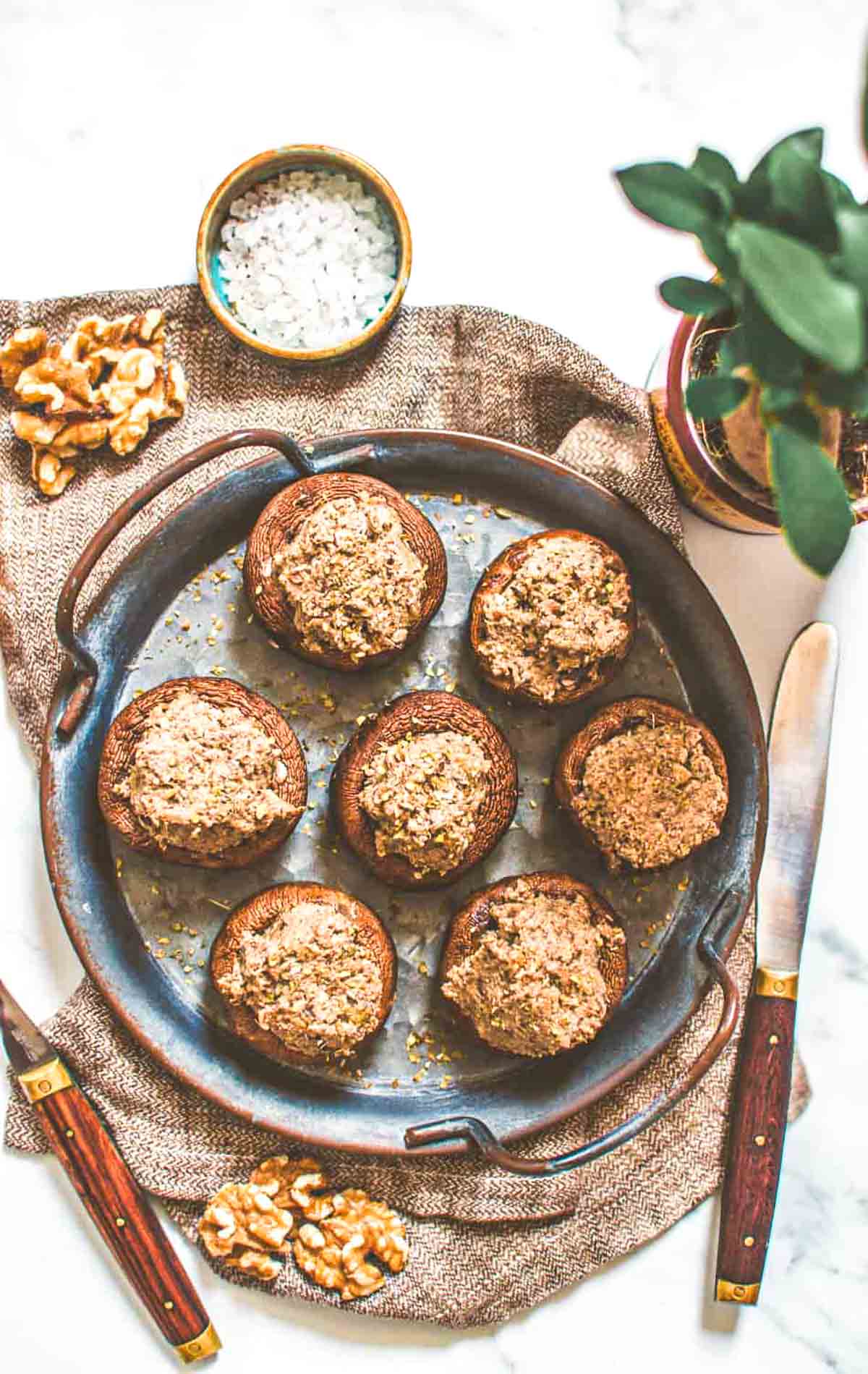 A metal platter filled with stuffed mushrooms, surrounded by walnuts, salt, and a plant in the background for a cozy, inviting feel.