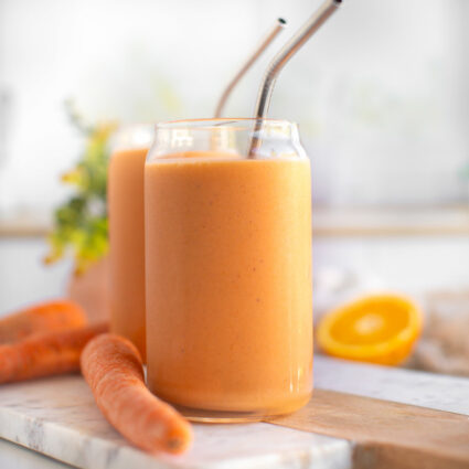 Carrot smoothies in glass cups with metal straws, next to carrots.