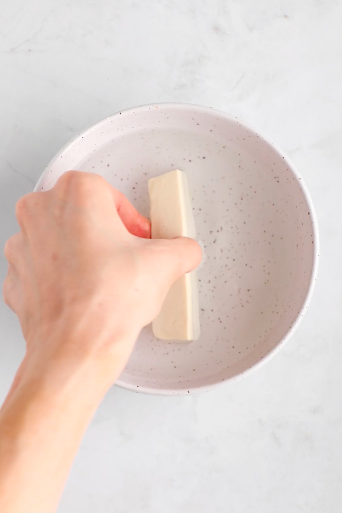 A hand dipping a slice of tofu into a bowl of water.