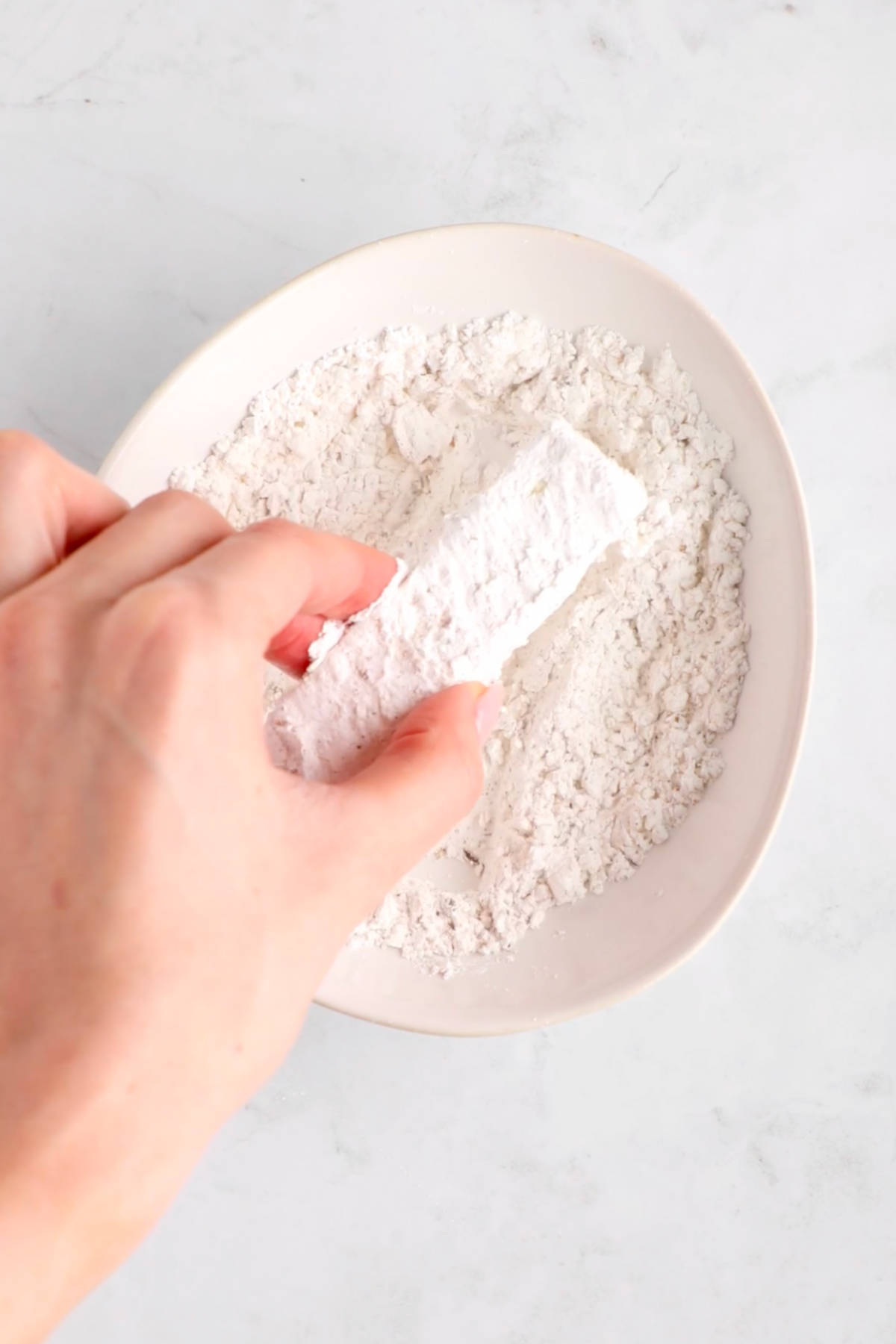 A hand rolling a slice of tofu in a bowl of cornstarch.