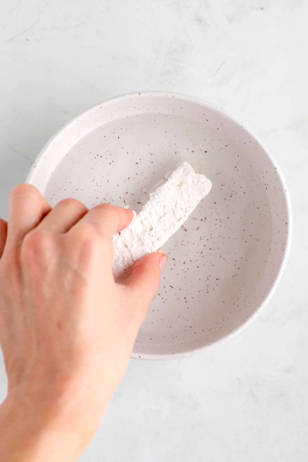 A hand dipping the cornstarch-coated tofu slice back into water.