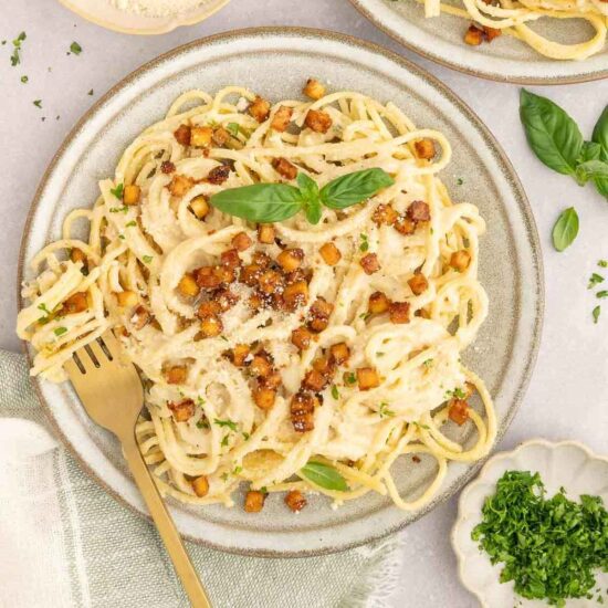 Creamy vegan carbonara topped with crispy tofu bacon, fresh herbs, and a basil garnish on a ceramic plate.