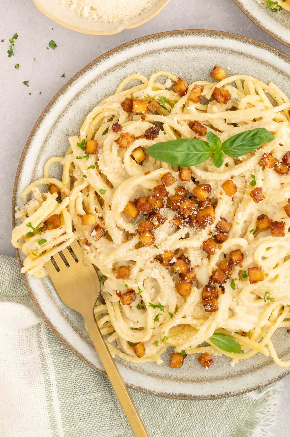 Plate of vegan carbonara topped with golden tofu bacon and fresh basil leaf.