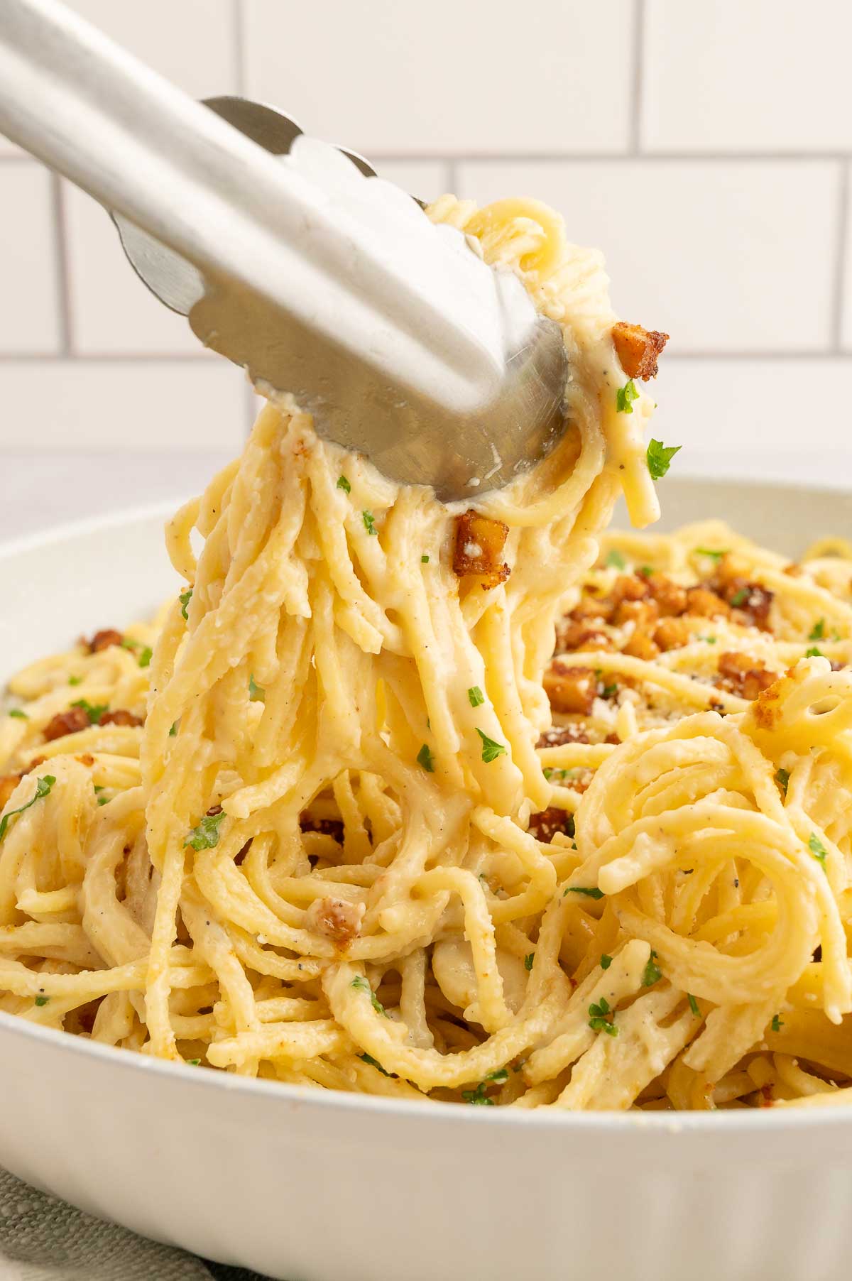 Close-up of vegan carbonara being lifted with tongs, showing creamy noodles and tofu bacon.
