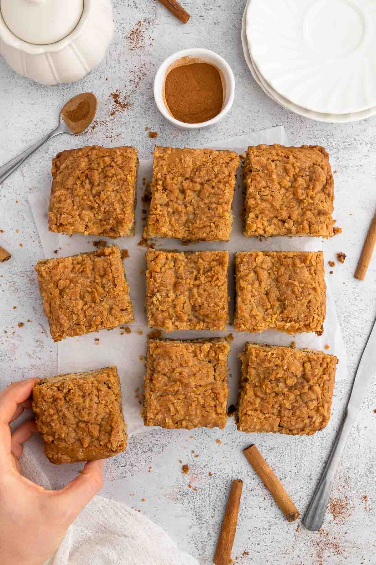 Vegan coffee cake sliced into nine squares on a piece of parchment paper along with sticks of cinnamon. 