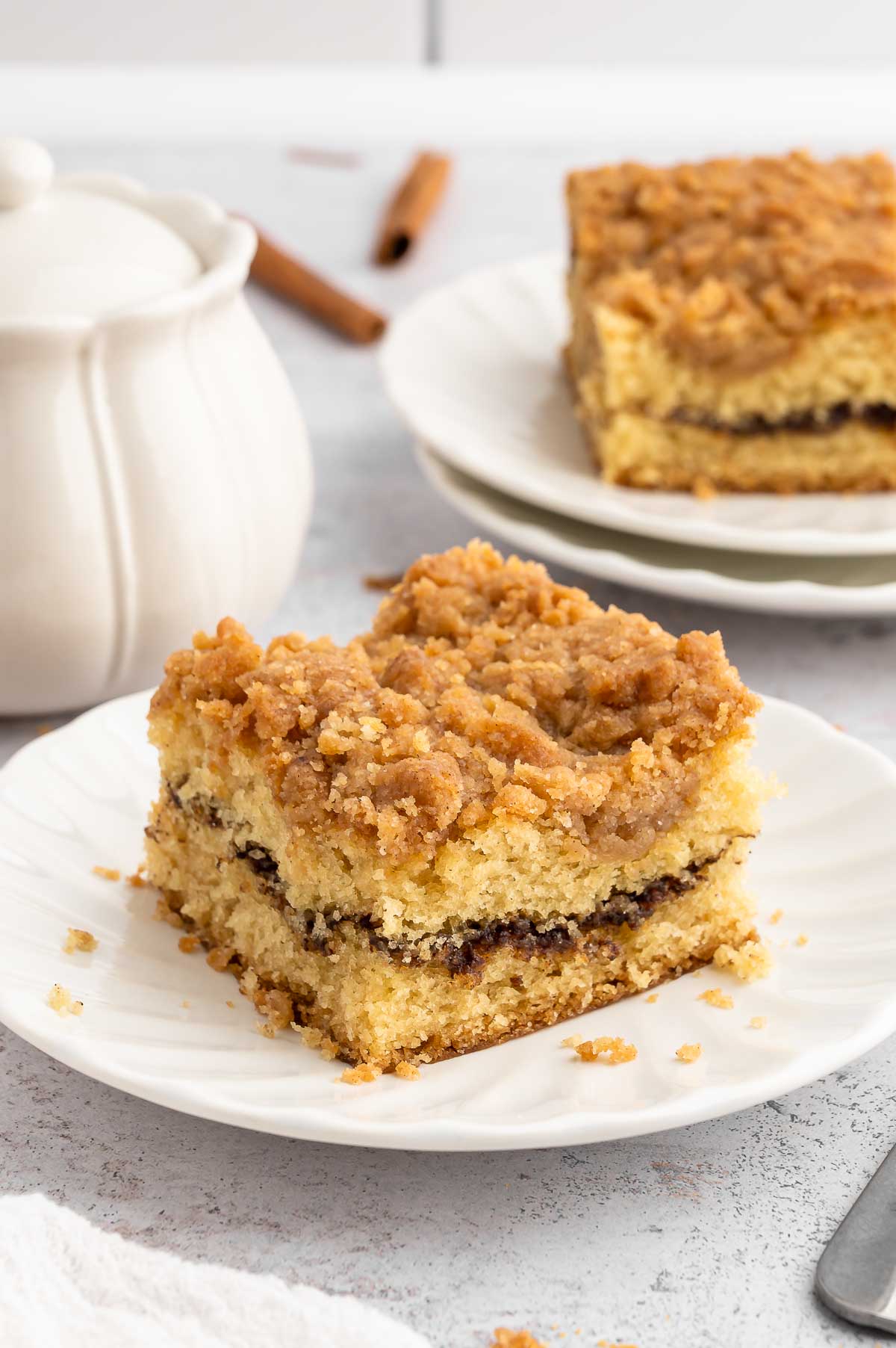 A piece of vegan coffee cake on a white plate next to a white pot of sugar with lid. 