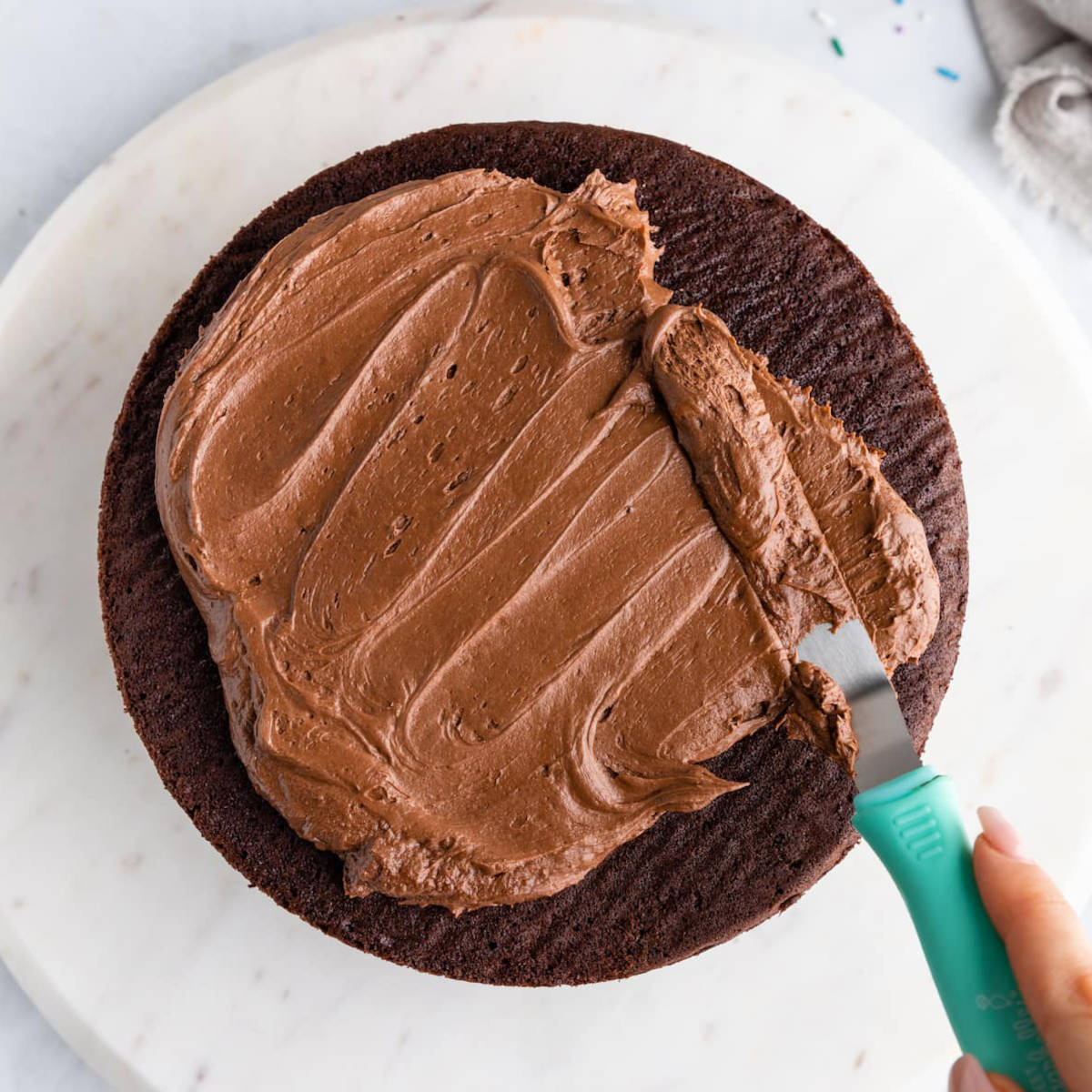 A hand spreading vegan chocolate frosting over a cake with an icing spatula.