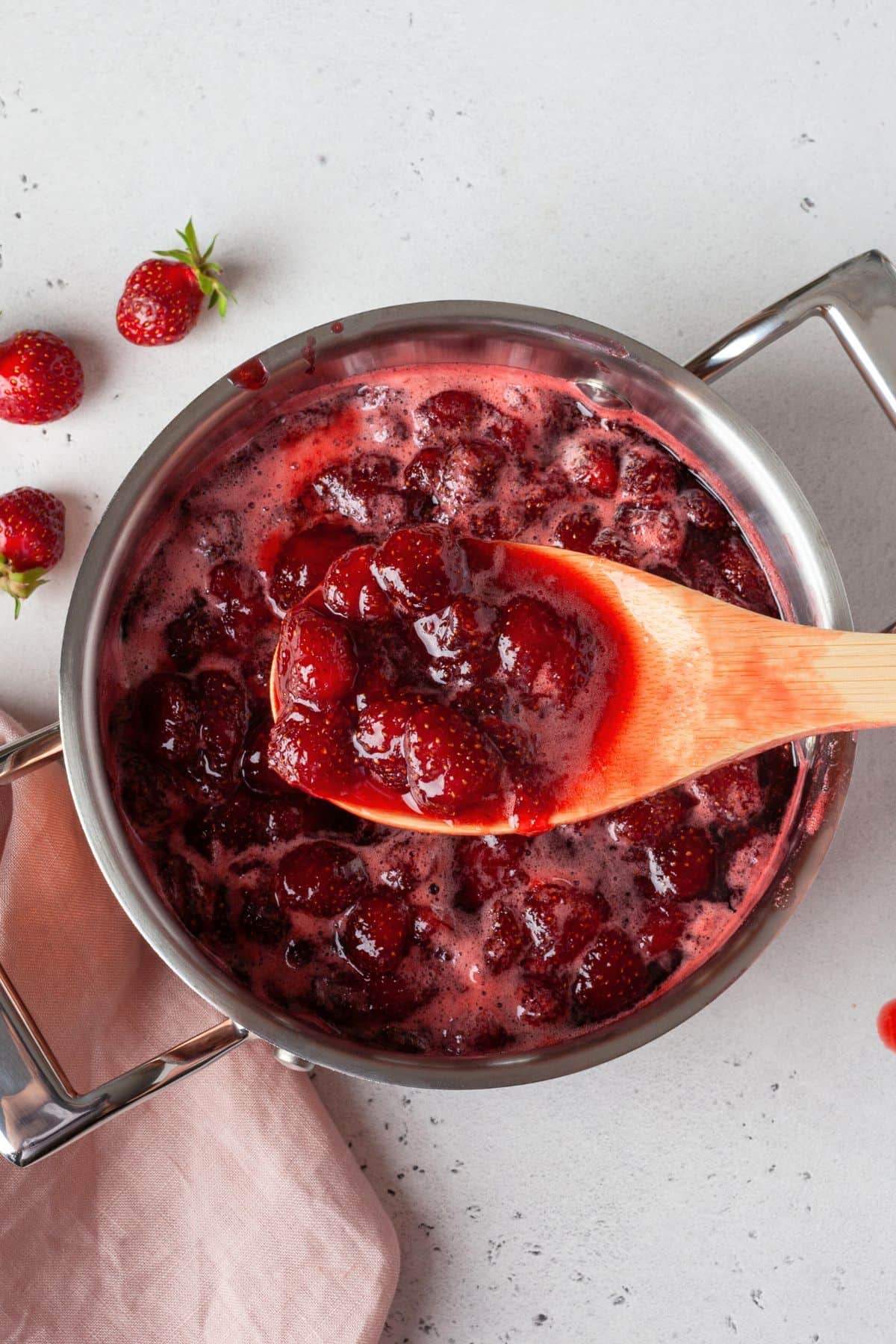 Vegan strawberry jam simmering in a pot with a wooden spoon scooping some up.