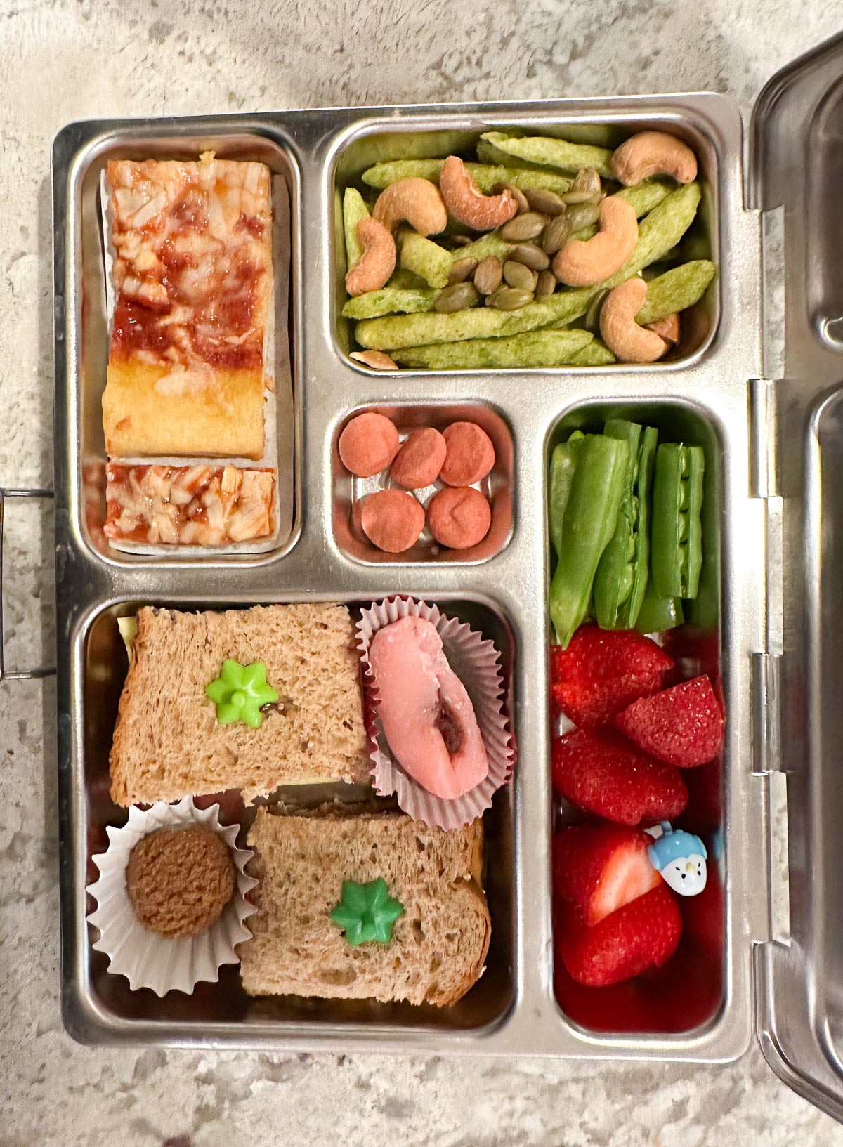 Colorful vegan lunchbox with mini pizza, fresh fruit, PB&J, and crunchy snacks.