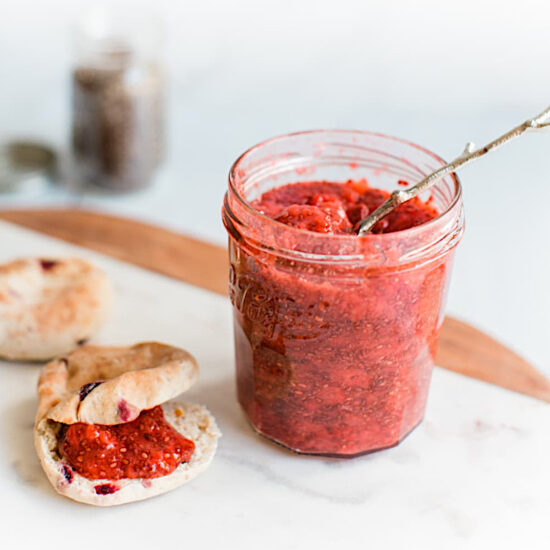 A jar of vegan strawberry jam next to a bagel smeared with strawberry jam.