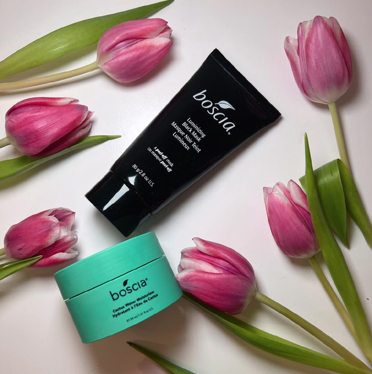 A black tube of Boscia mask and a green jar of Boscia face moisturizer on a white background with pink tulips.