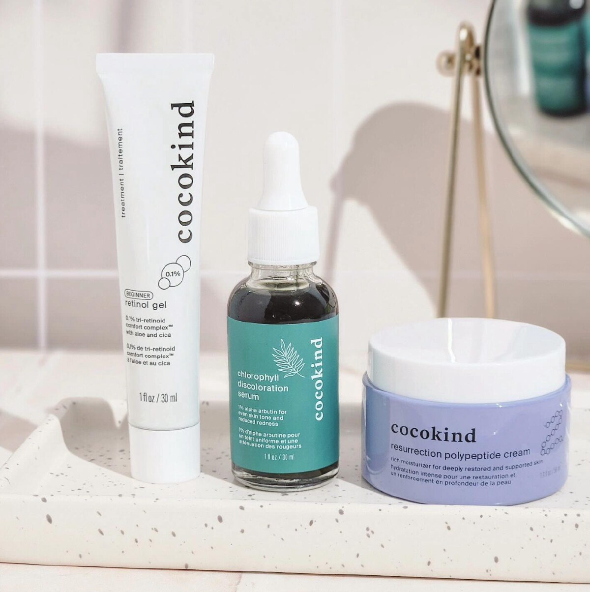 A tube, a bottle and a jar of Cocokind skincare in a marble-like dish against a bathroom tile background.