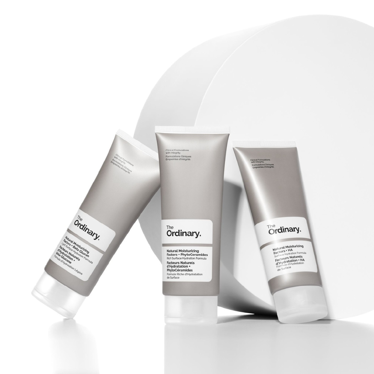 Three tubes of silver and white face lotion by The Ordinary against a white background with white, solid, circular standing shape.