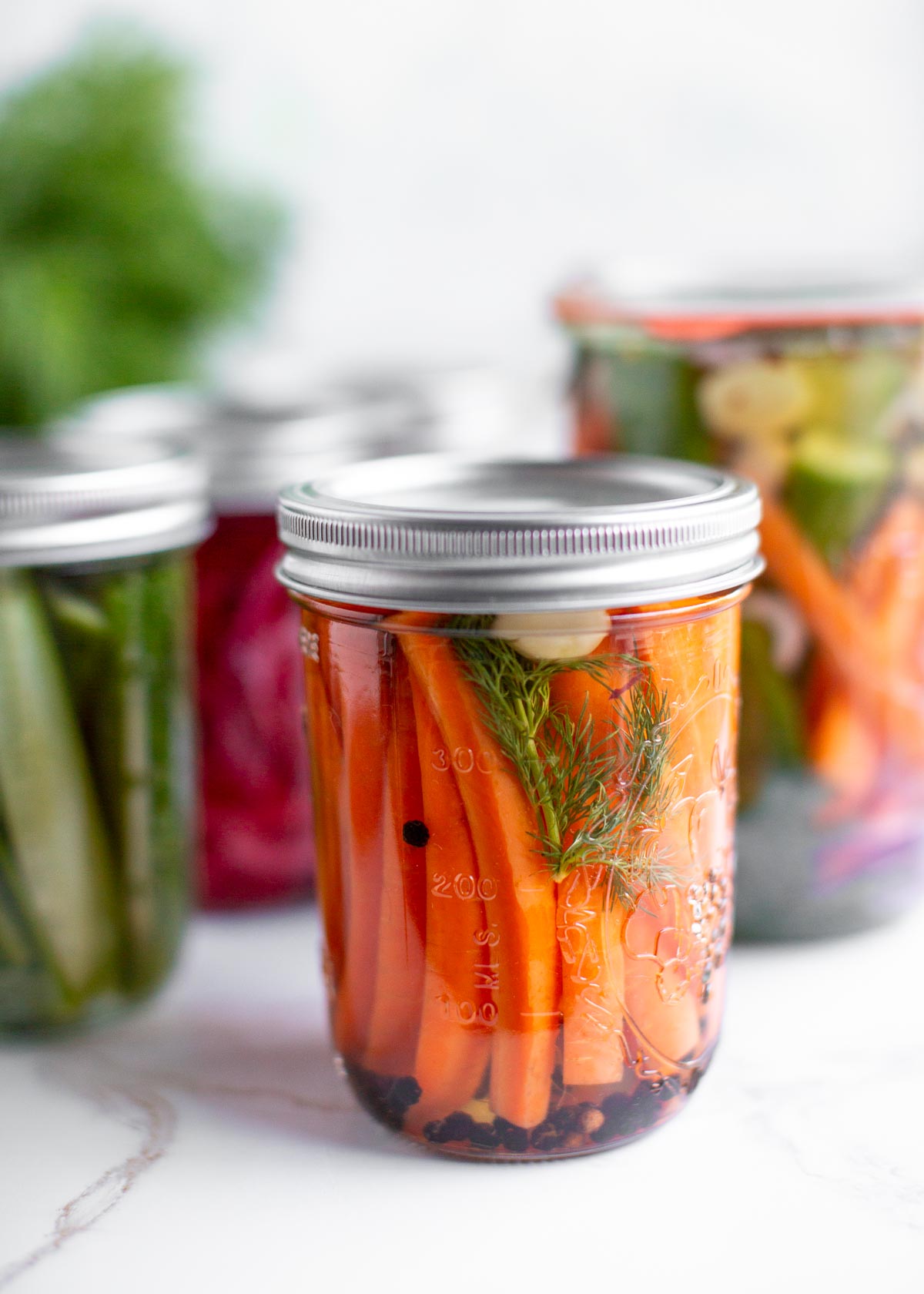 Pickled carrots in a glass jar with various jars of pickled vegetables behind it.