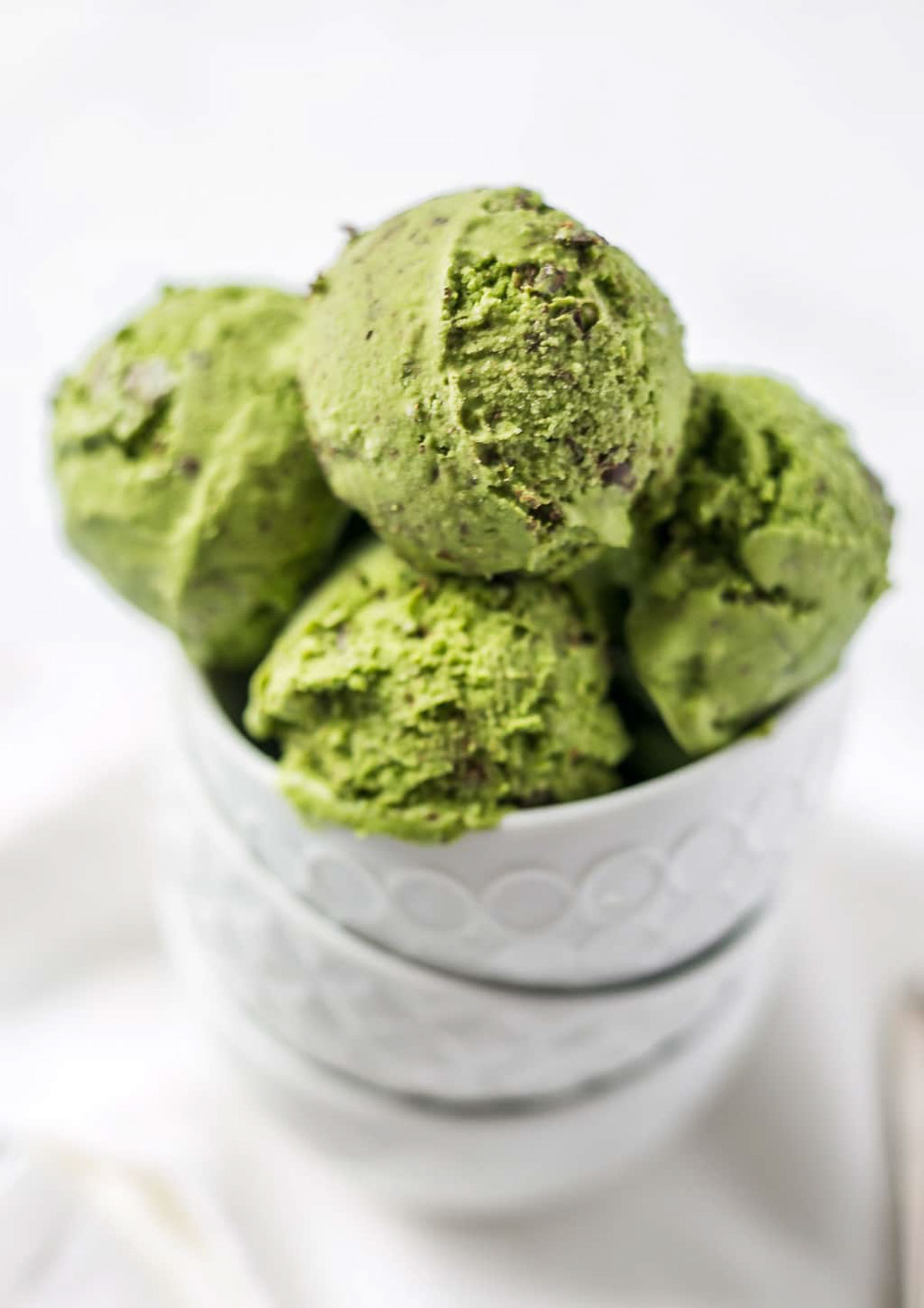 Scoops of vegan matcha ice cream stacked in a white bowl, showing creamy texture and chocolate chips.
