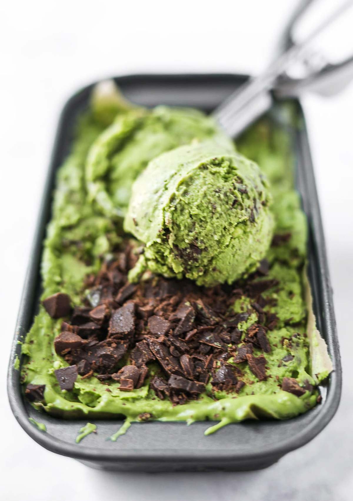 Close-up of a scoop of vegan matcha ice cream being served from a container, topped with vegan chocolate chips.
