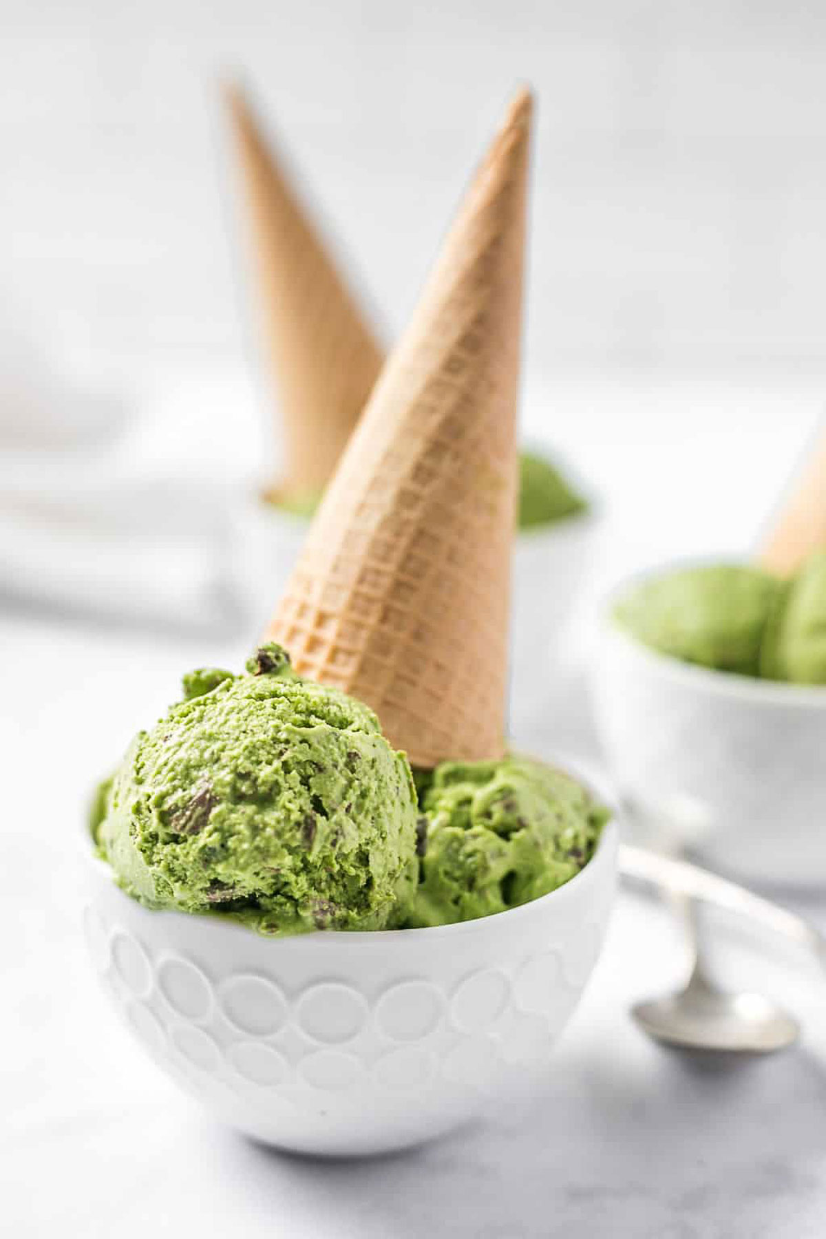 Scoops of vegan matcha ice cream in white bowls topped playfully with upside-down waffle cones.