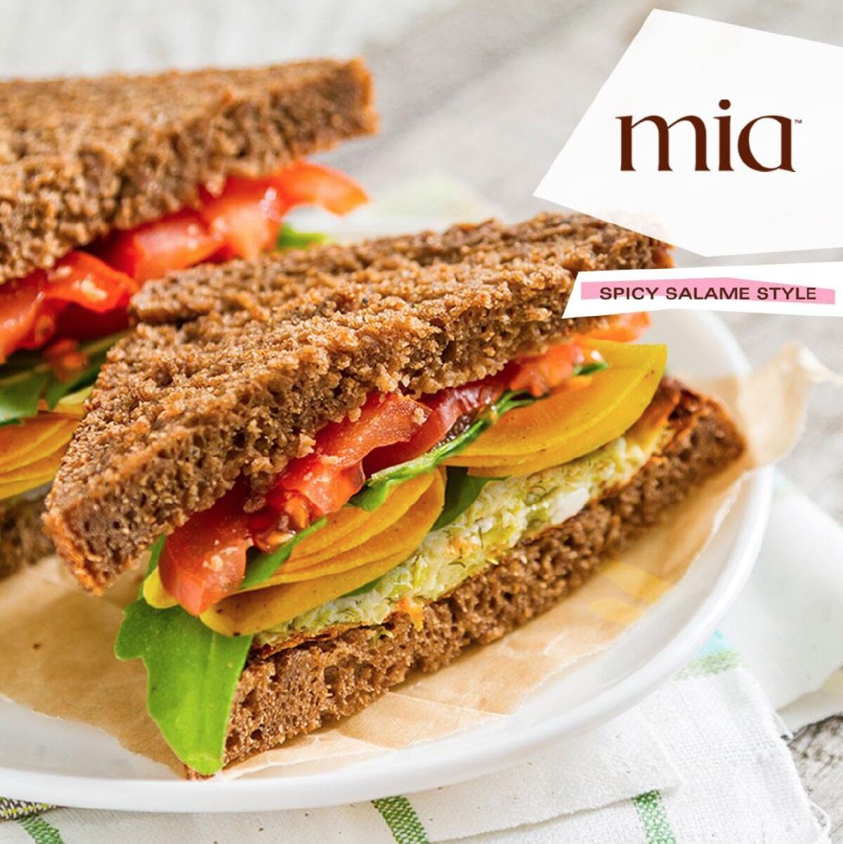 Two triangle slices of sandwich containing Mia Foods vegan deli slices on a white plate on top of a striped napkin.