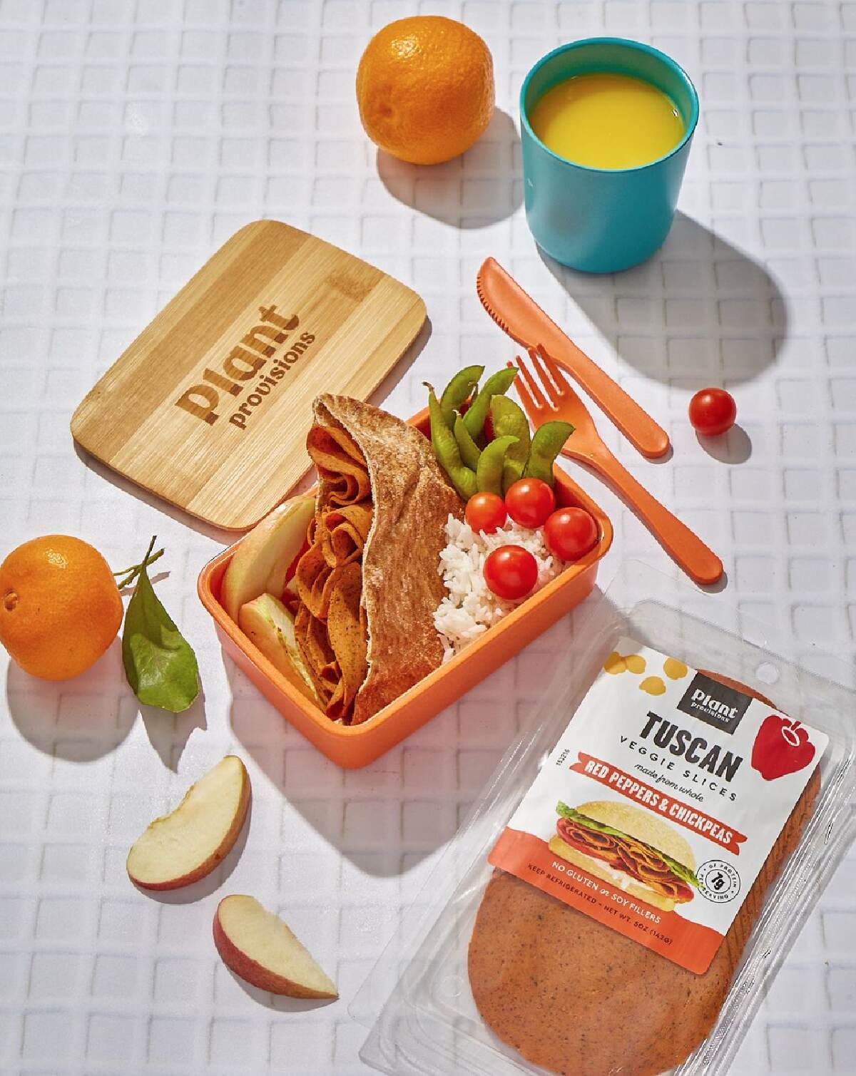 An orange lunch box with bamboo lid and a package of Plant Provisions Tuscan vegan deli slices with a glass of juice and fruit. 