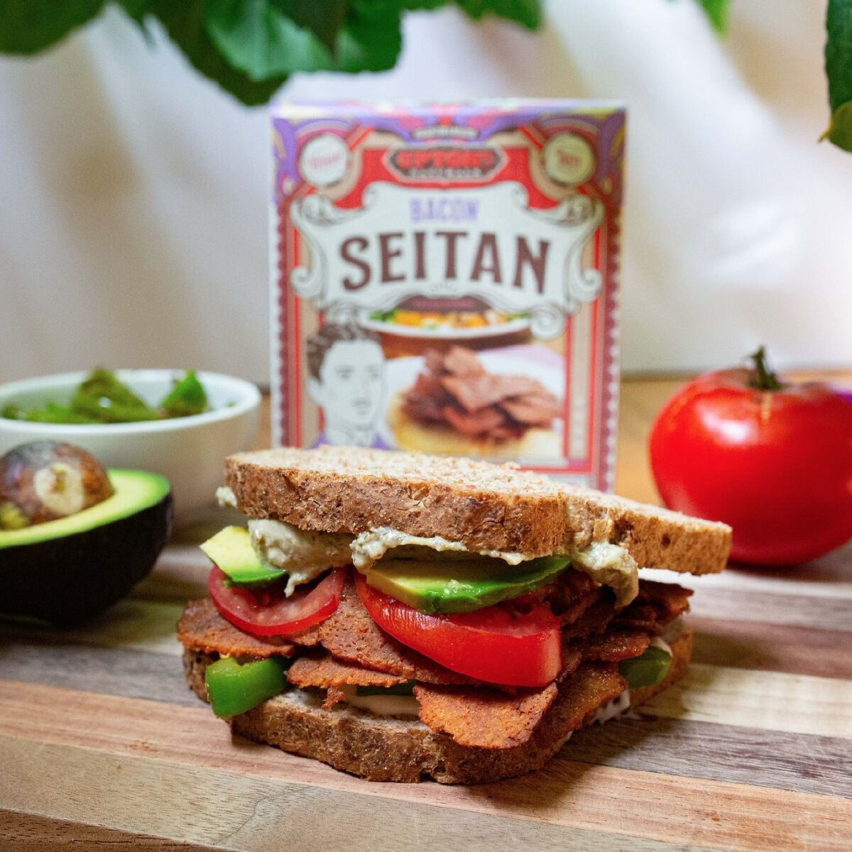 Uptons Naturals seitan bacon box, tomato, and BLT sandwich on a wooden table.