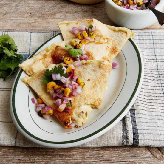 Three vegan quesadillas on a plate topped with diced red onion, corn kernels, and cilantro leaves.