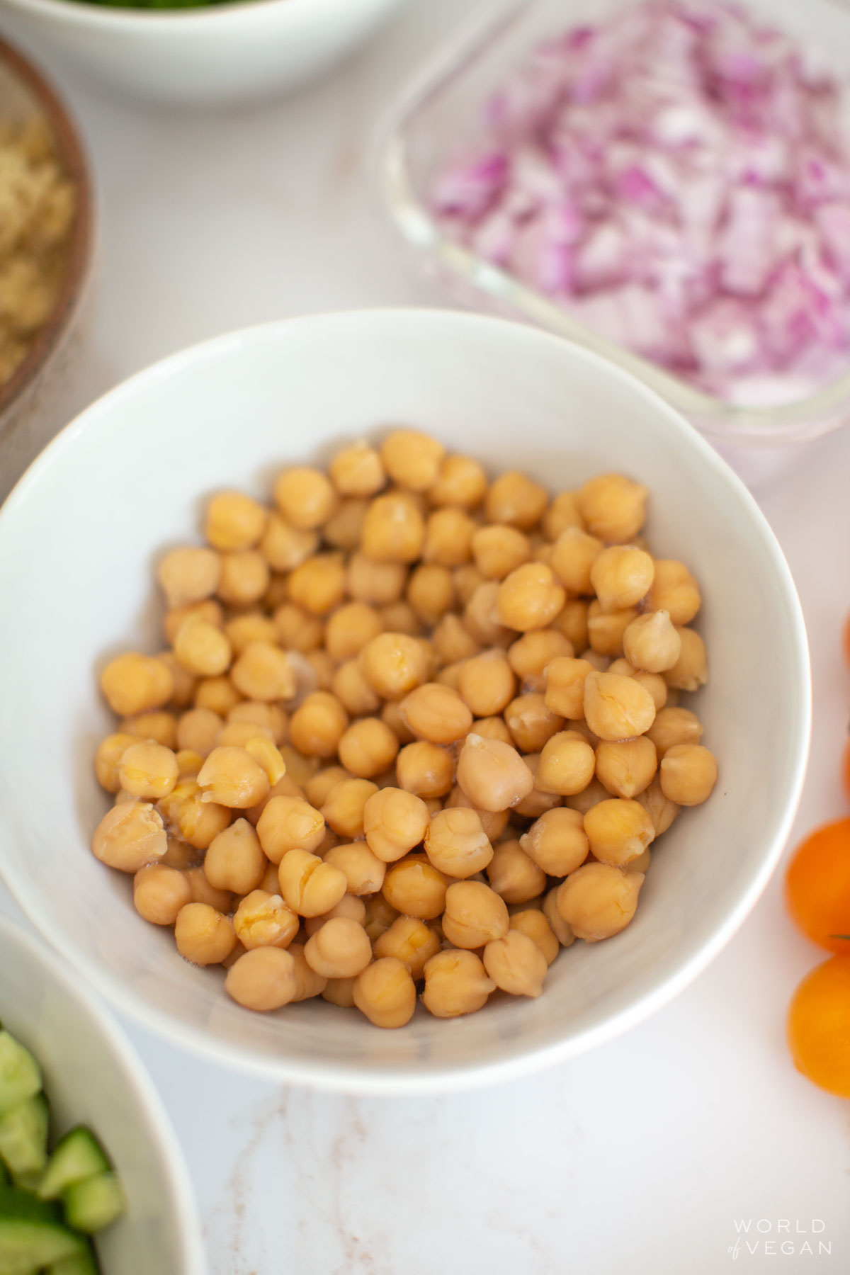 A bowl of chickpeas surrounded by bowls of various vegetables like red onion, cauliflower, and cucumber.