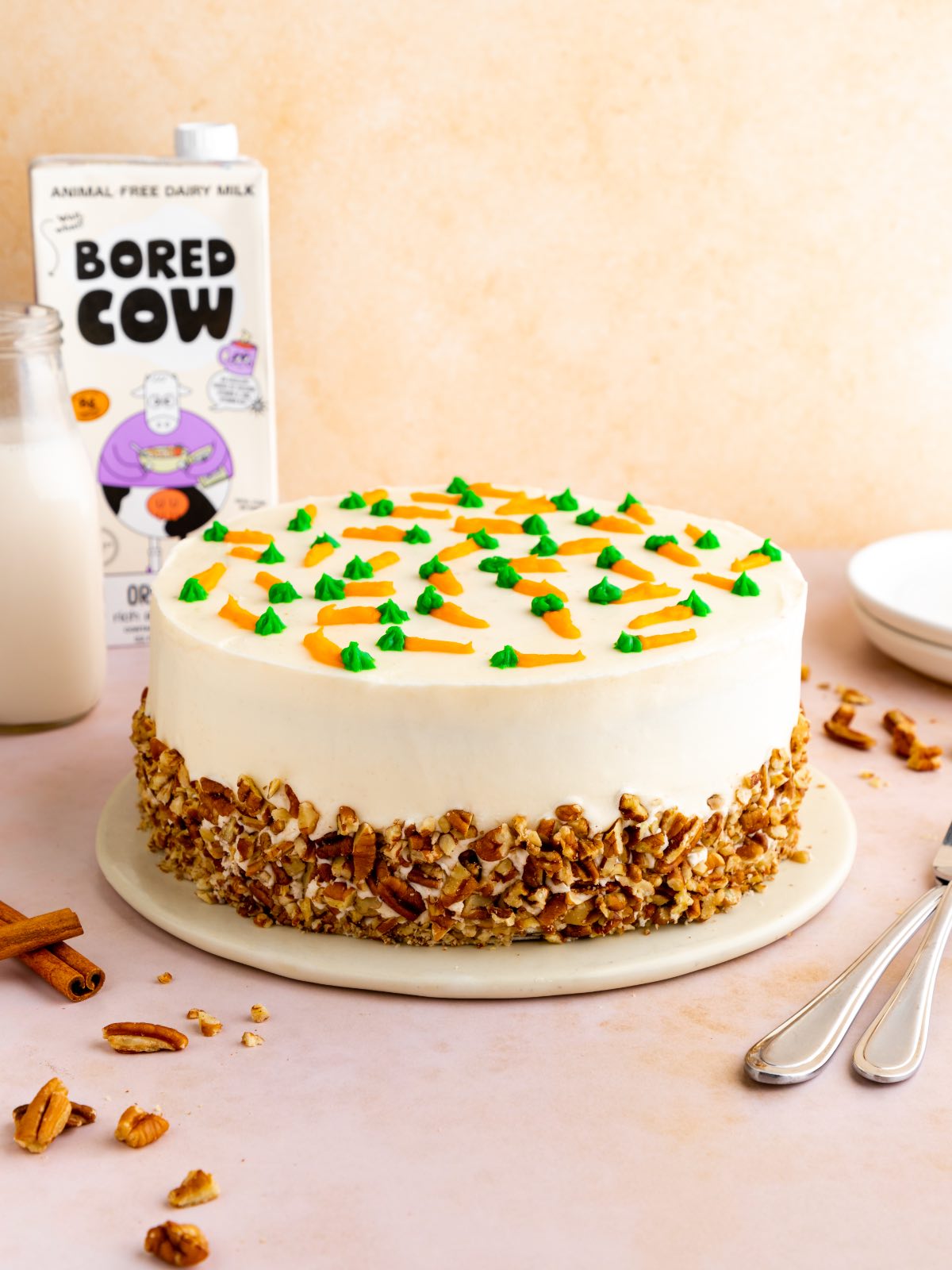 A whole beautifully decorated vegan carrot cake topped with cream cheese frosting and little frosted carrots and chopped nuts around the outside. 