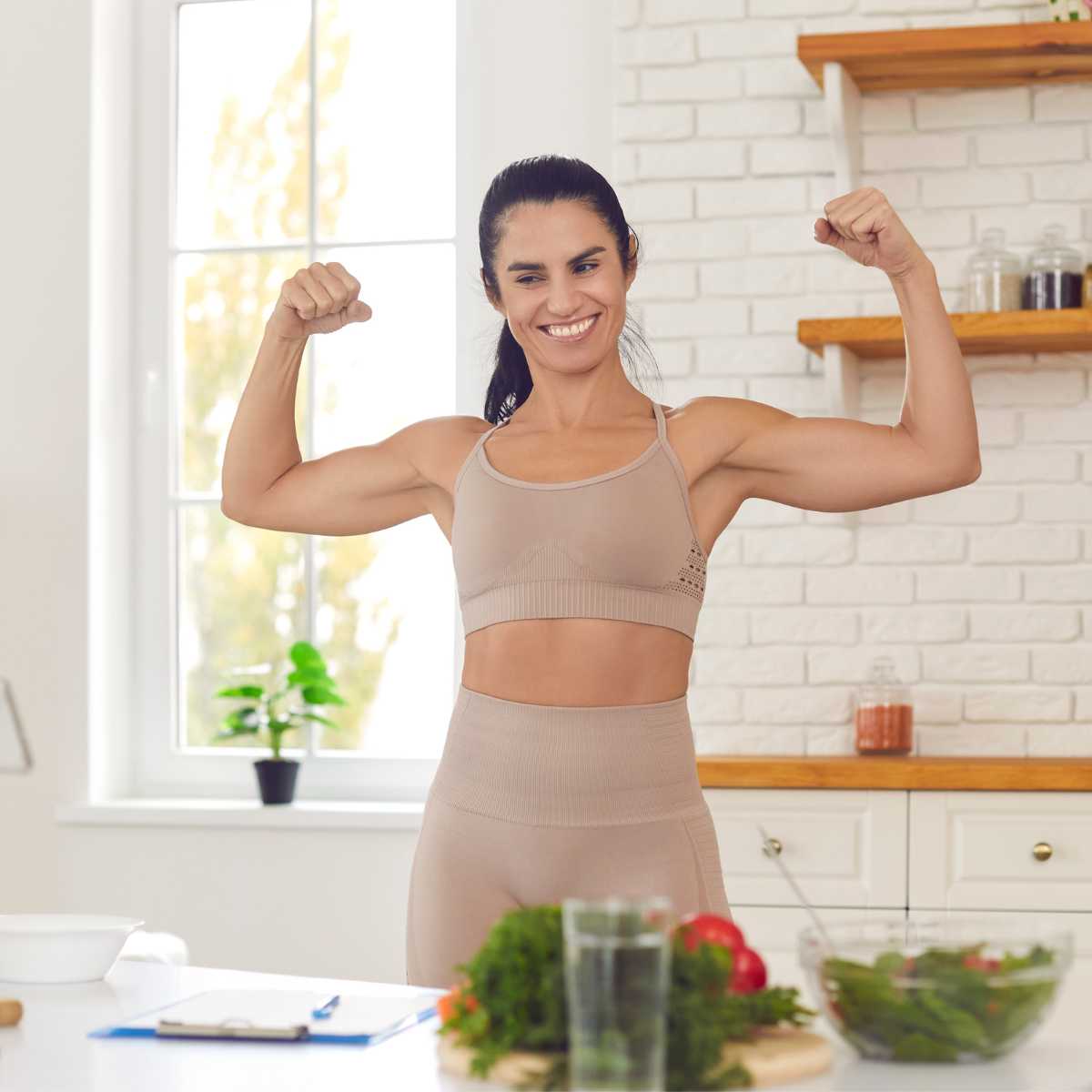 Woman flexing her muscles in fitness gear in her kitchen with vegan pre-workout food and water around her.