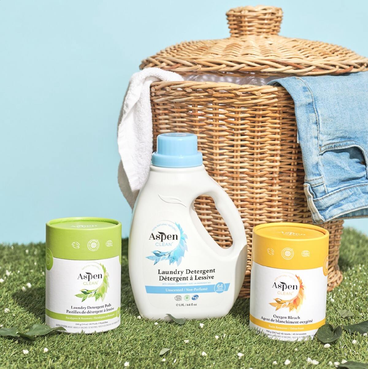 Two types of Aspen Clean laundry detergent and one container of oxygen bleach on a faux grassy spot next to a wicker basket full of laundry.