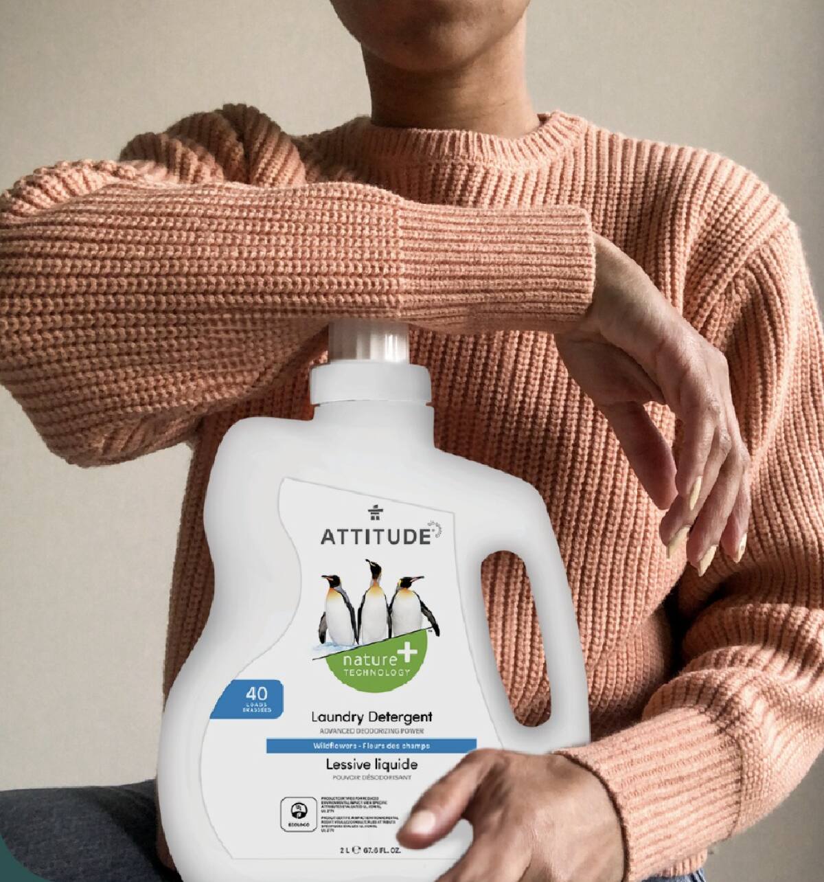 A woman with a beige pink sweater leaning on a white plastic bottle of Attitude Living Laundry Detergent.
