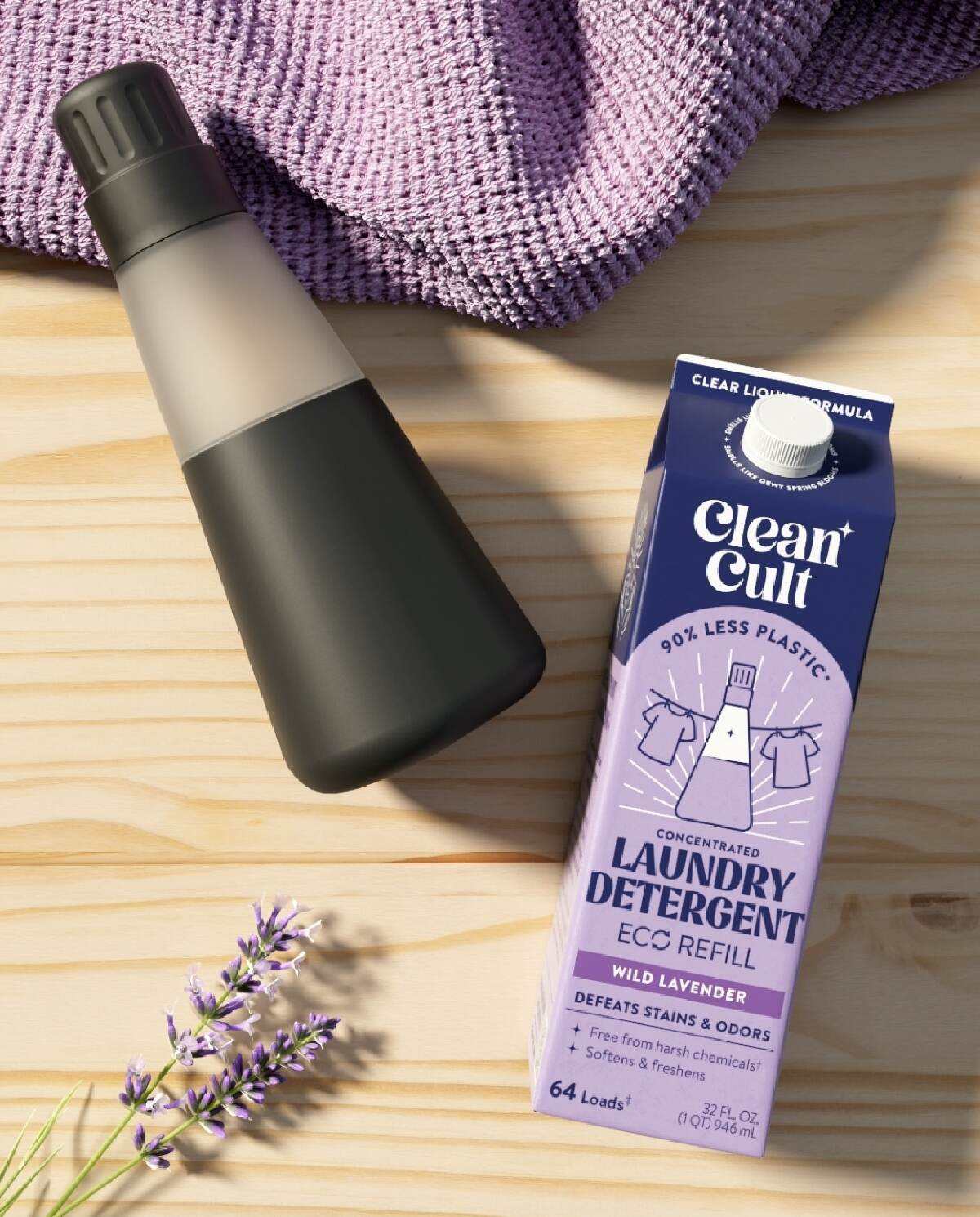 A clear and black plastic reusable bottle next to a cardboard refill of Clean Cult laundry laundry detergent next to a purple towel on a wood background. 