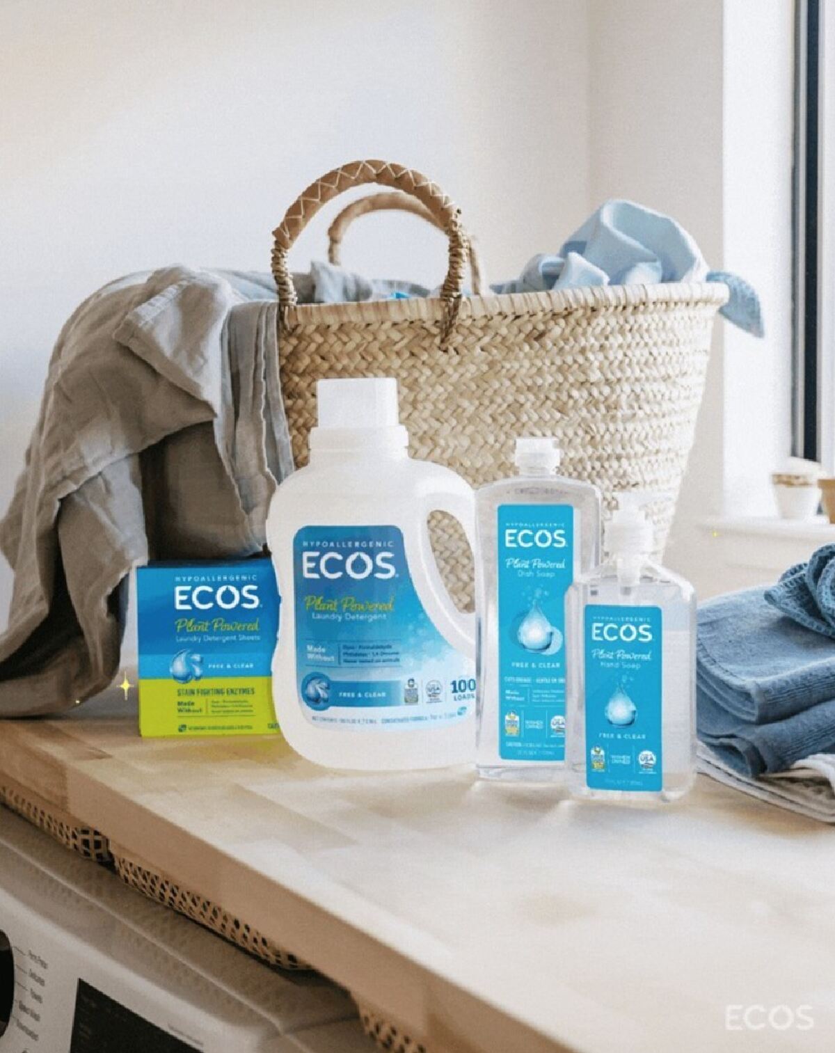 Ecos plant-powered laundry detergent, laundry sheets and other cleaning products in front of a straw bag filled with laundry on a wood topped surface next to a bright window.