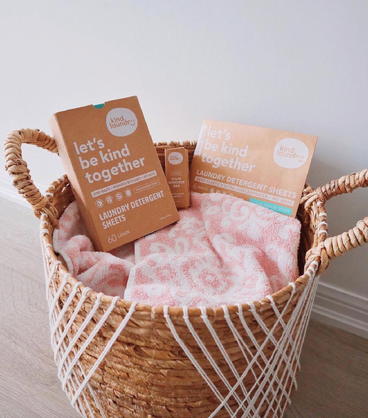 Laundry detergent sheets and stain bar by Kind Laundry in cardboard boxes in a straw laundry basket with pink towels. 