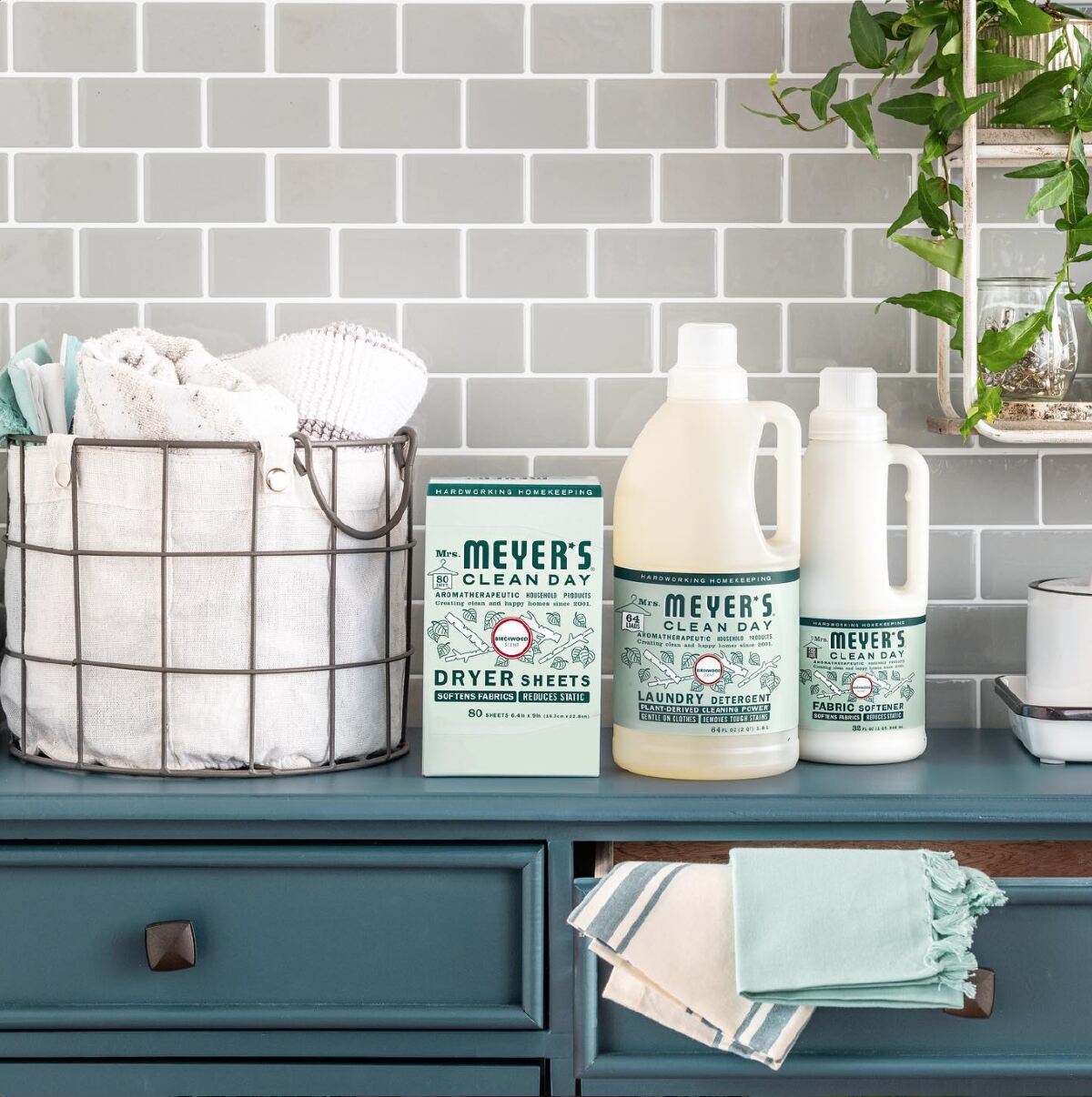 Mrs. Meyers Clean Day dryer sheets, laundry detergent and fabric softener on a dark teal desk next to a metal basket full of white towels against a gray tile background.