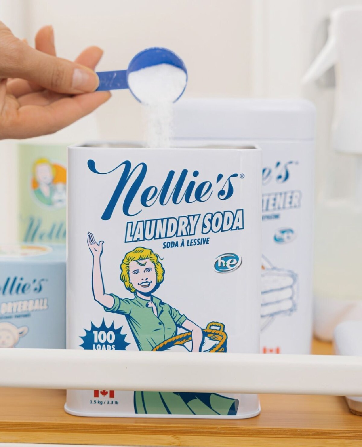 A white and blue metal container of Nellie's Laundry Soda shown with a hand using a blue scoop to pour it in.