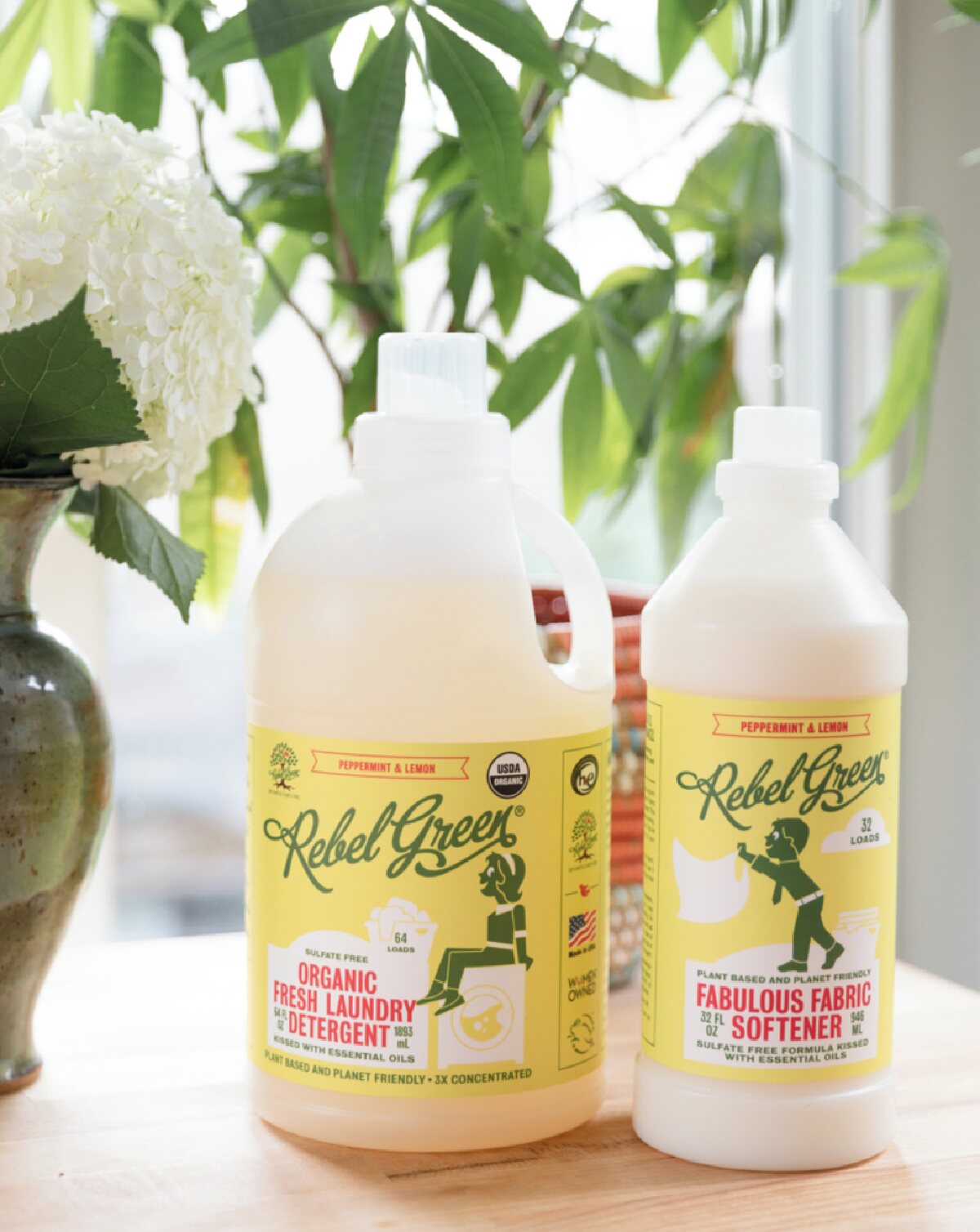 Two clear plastic bottles with yellow labels of Rebel Green's organic laundry detergent and fabric softener on a wooden table next to white flowers and plants.